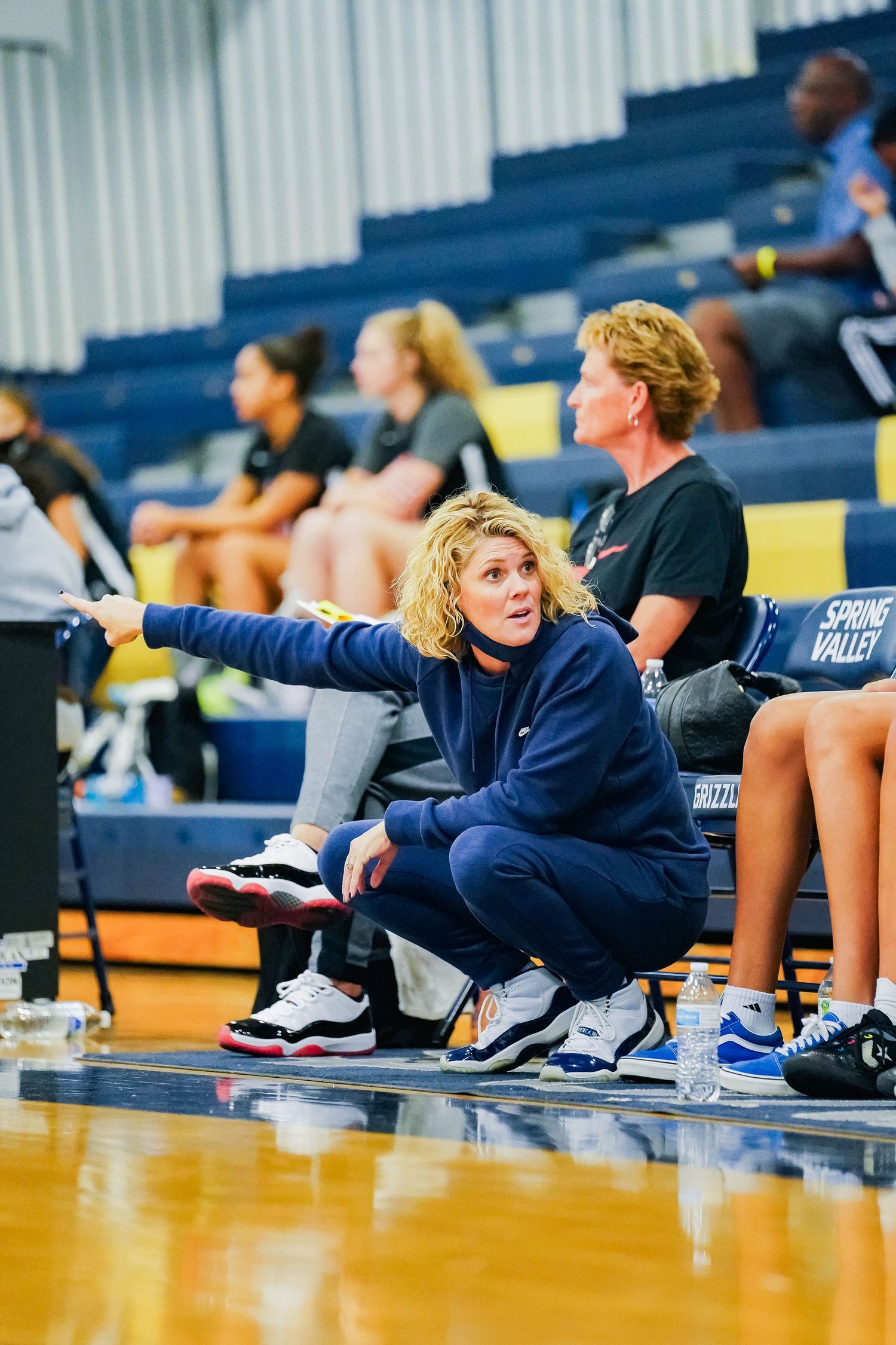 Coach gestures from sideline during basketball game wearing navy sweatshirt.