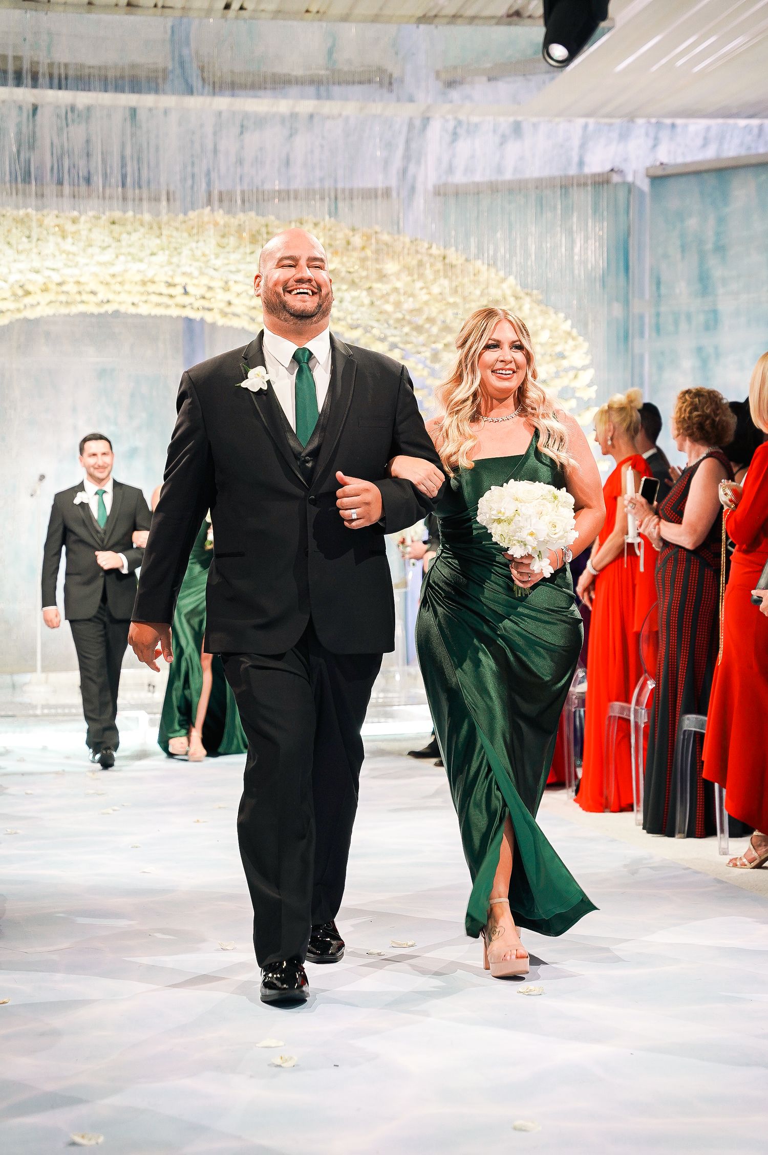 Wedding party members walk down the aisle in emerald green and red formal attire.