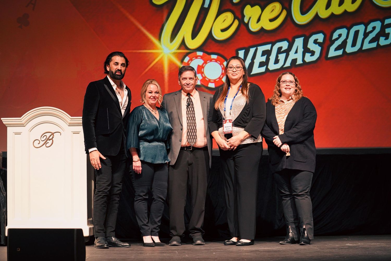 People in formal attire gathered on stage at a Vegas event with red lighting and signage.