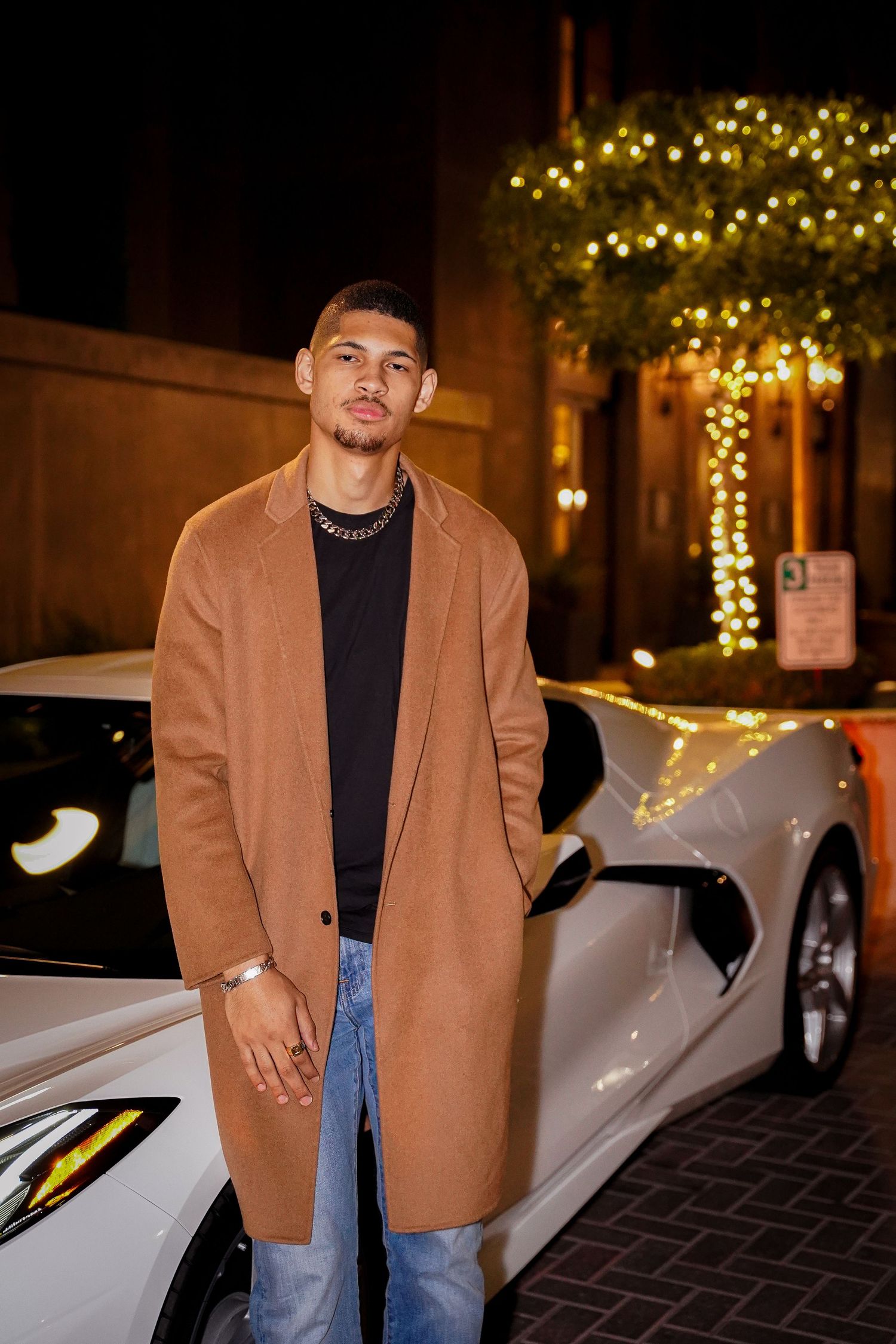 Person in camel coat and black turtleneck poses next to white luxury car at night.