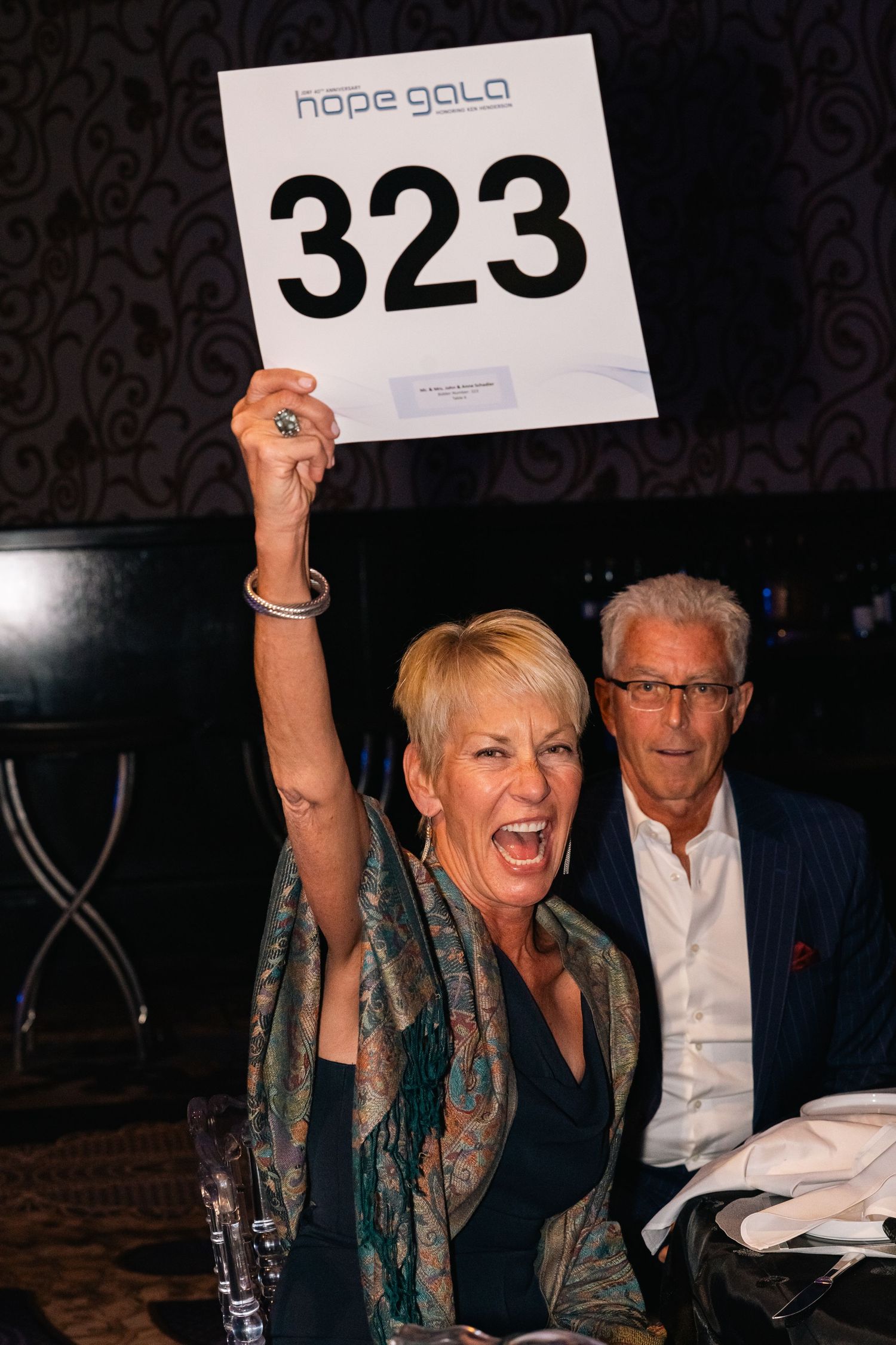 Person at gala event enthusiastically holding up auction paddle number 323.