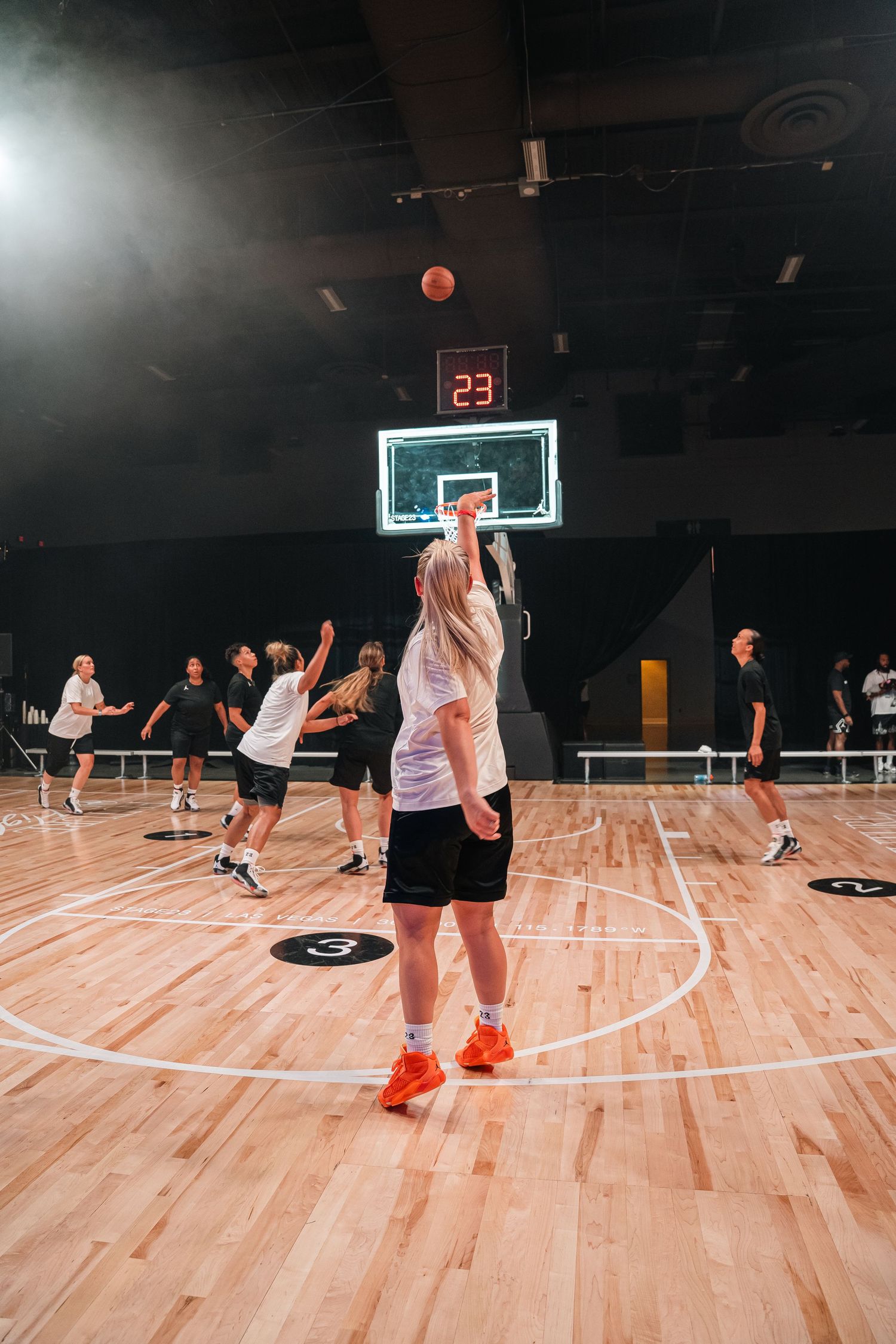 Basketball players practicing shots on an indoor court with a digital scoreboard displaying 23.