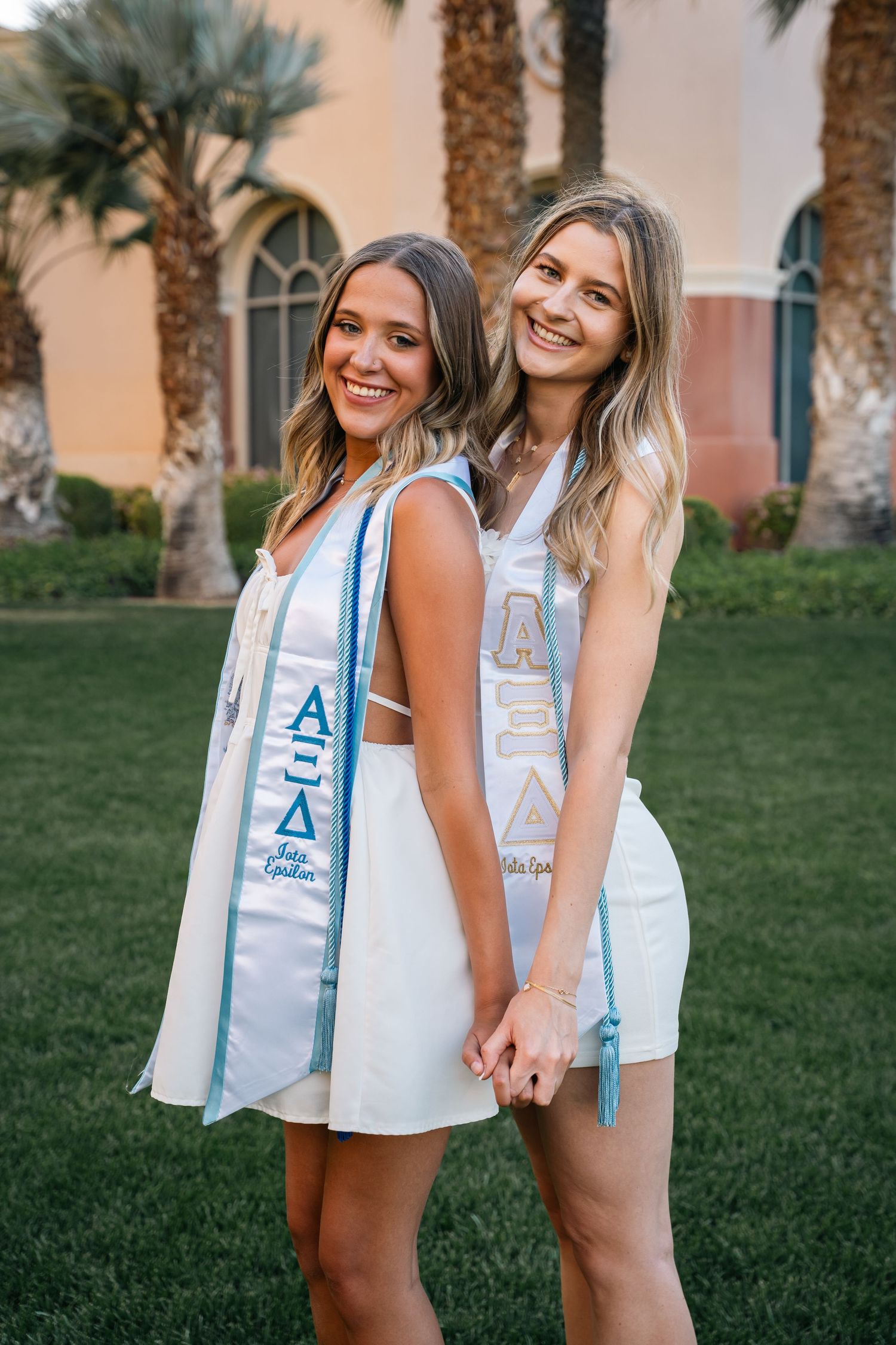 Two sorority sisters in white dresses share a joyful moment while wearing their graduation stoles.