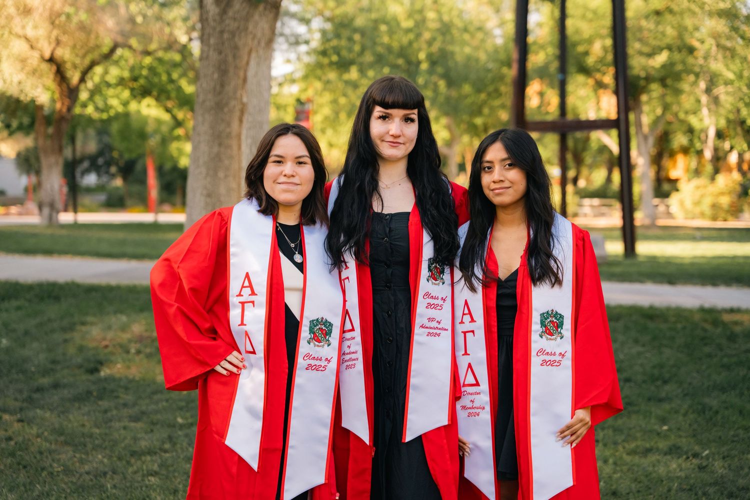 Three graduates in red graduation gowns and white stoles pose together outdoors on campus.