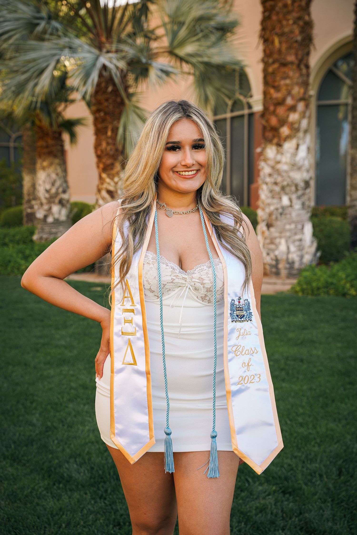 Graduation photos in white dress and decorative sash against palm tree backdrop.