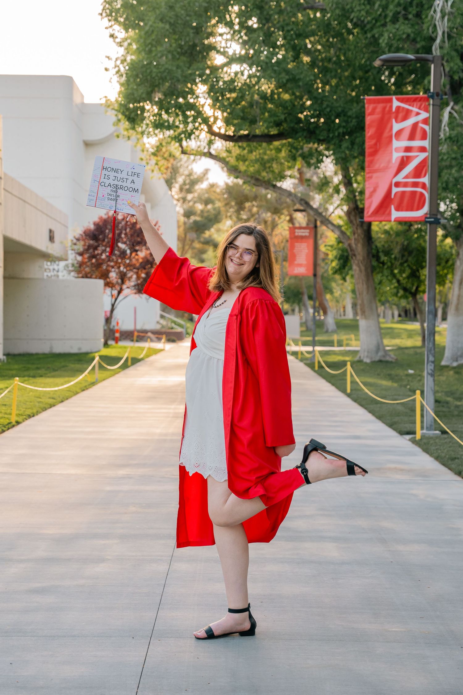 Graduate in red cap and gown poses playfully on campus pathway.