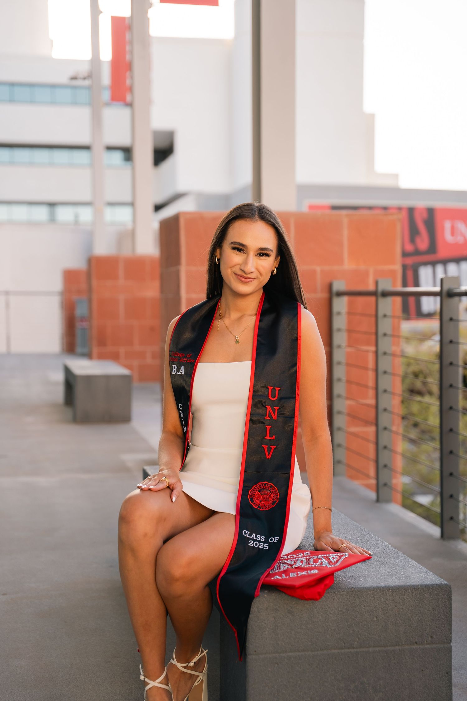 Graduate in formal attire sits on concrete ledge during outdoor photo session.