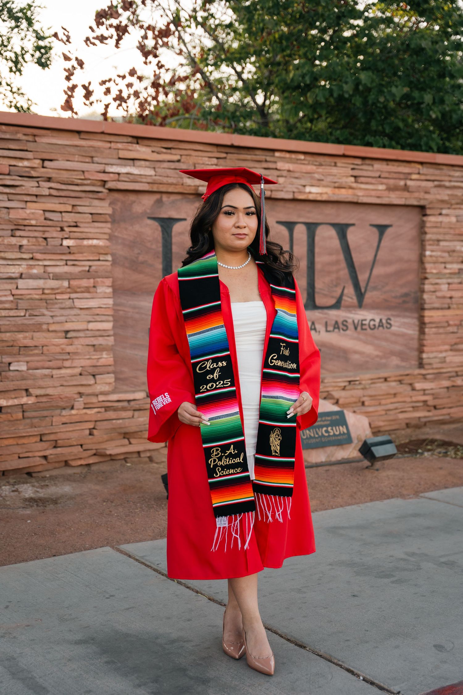 Graduate wearing red cap and gown displays cultural graduation stole and diploma.