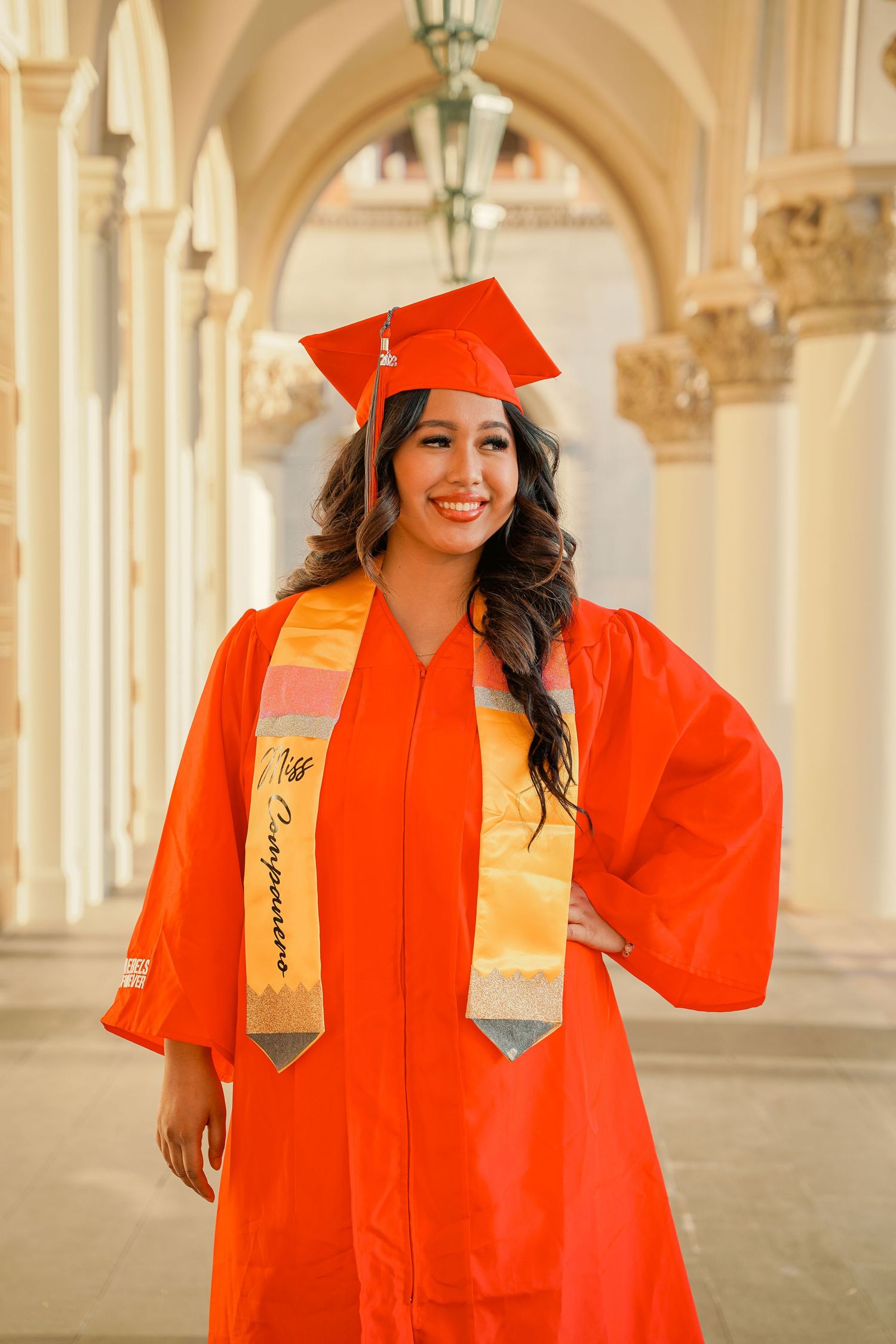 Graduate wearing bright orange cap and gown poses in elegant arched hallway with decorative columns.