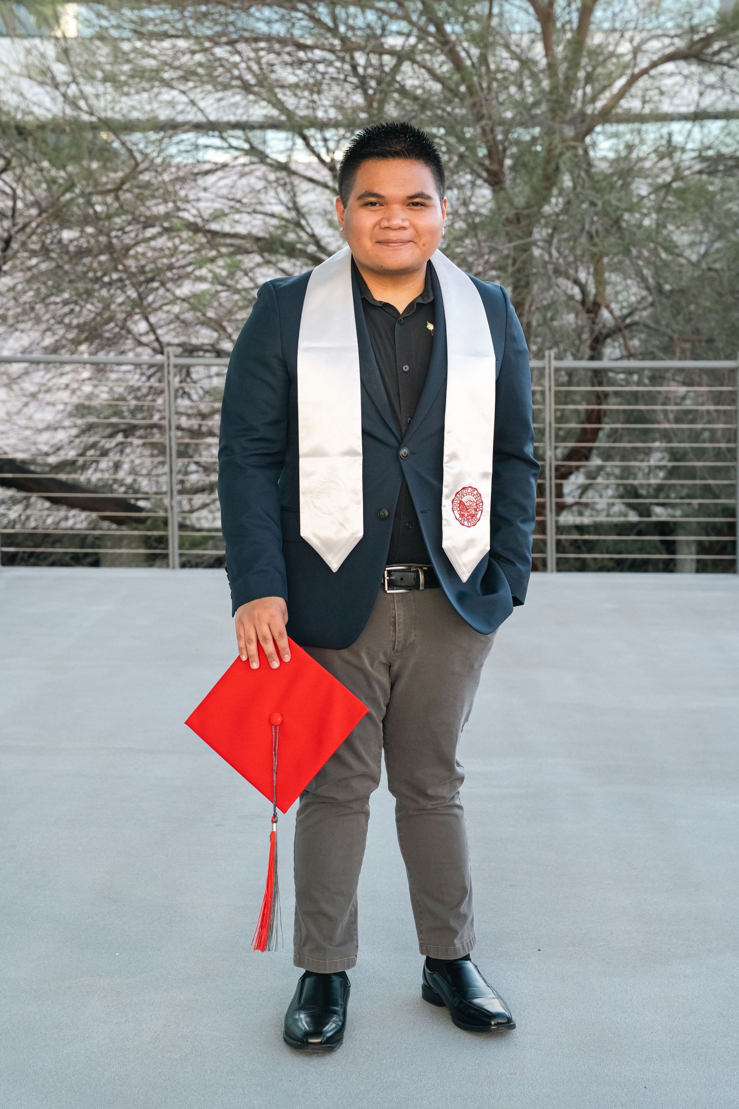 Graduate wearing navy blazer and white stole poses with red diploma cover.