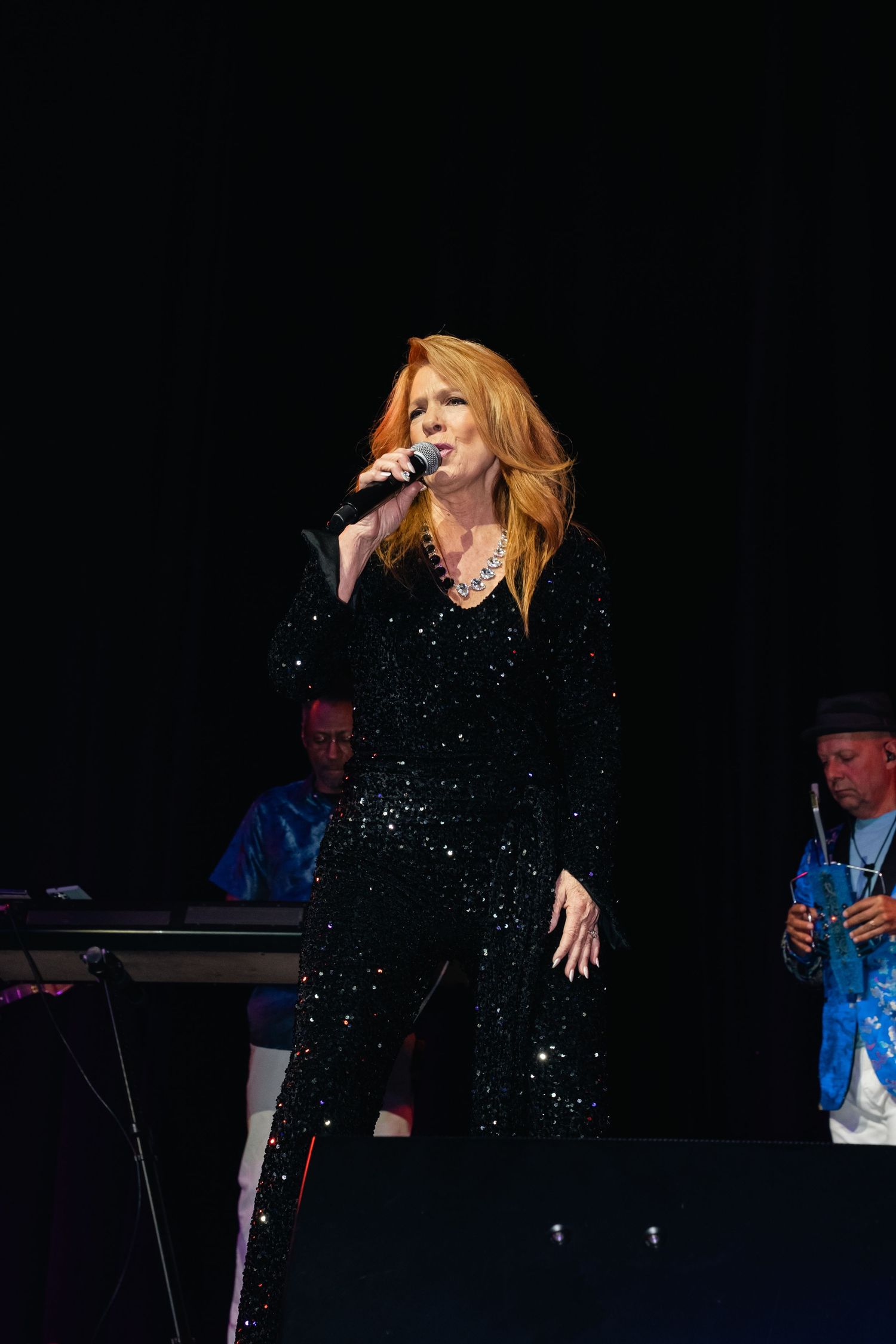 Performer in sparkling black outfit singing on stage with dramatic lighting.