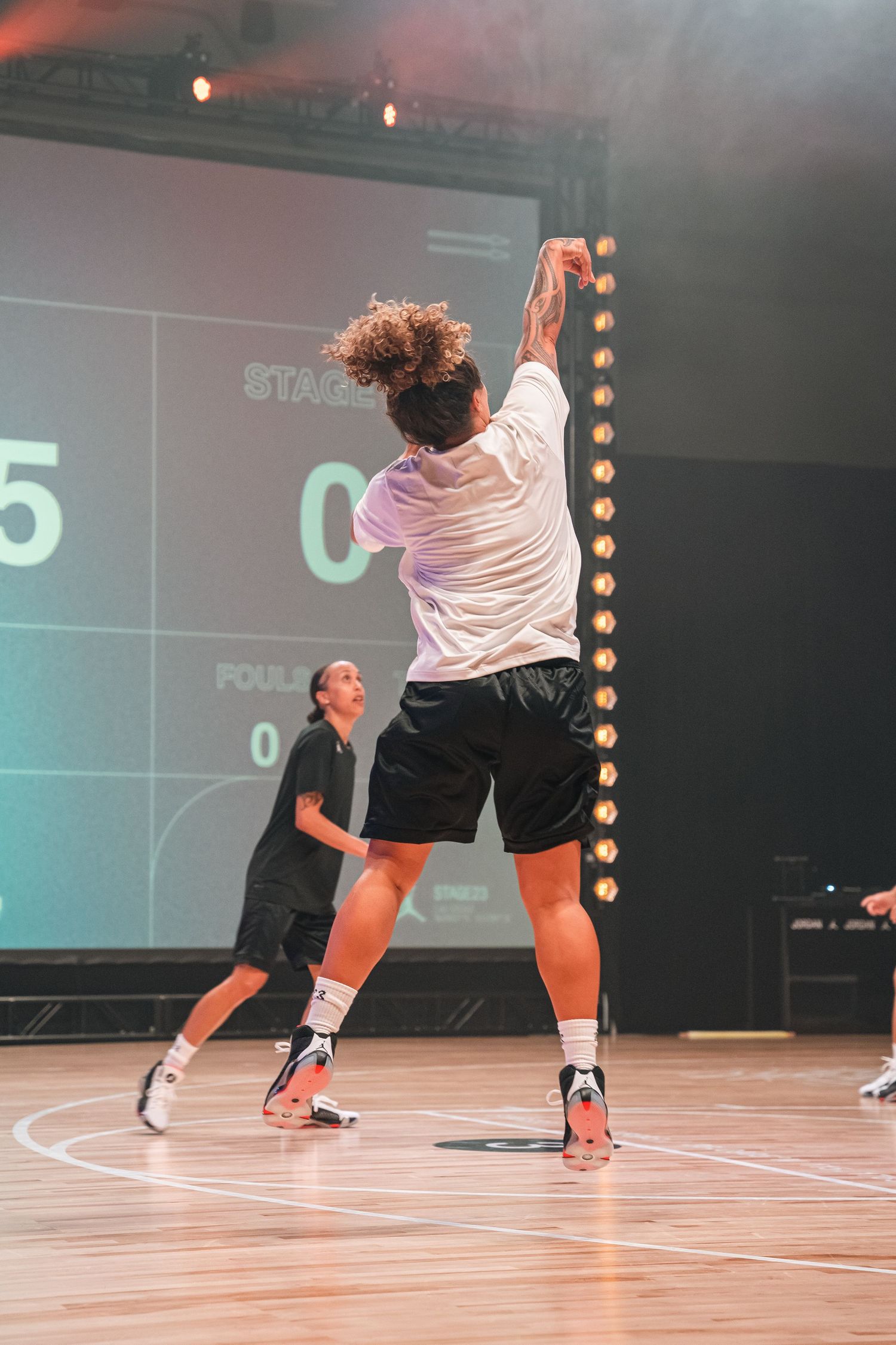 Player taking a jump shot on indoor basketball court with digital scoreboard display.