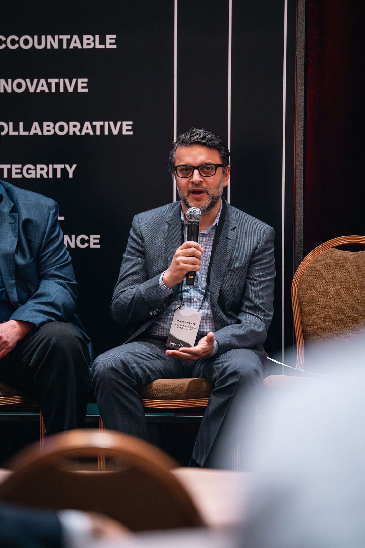Speaker in suit jacket with glasses holding microphone during conference panel discussion.