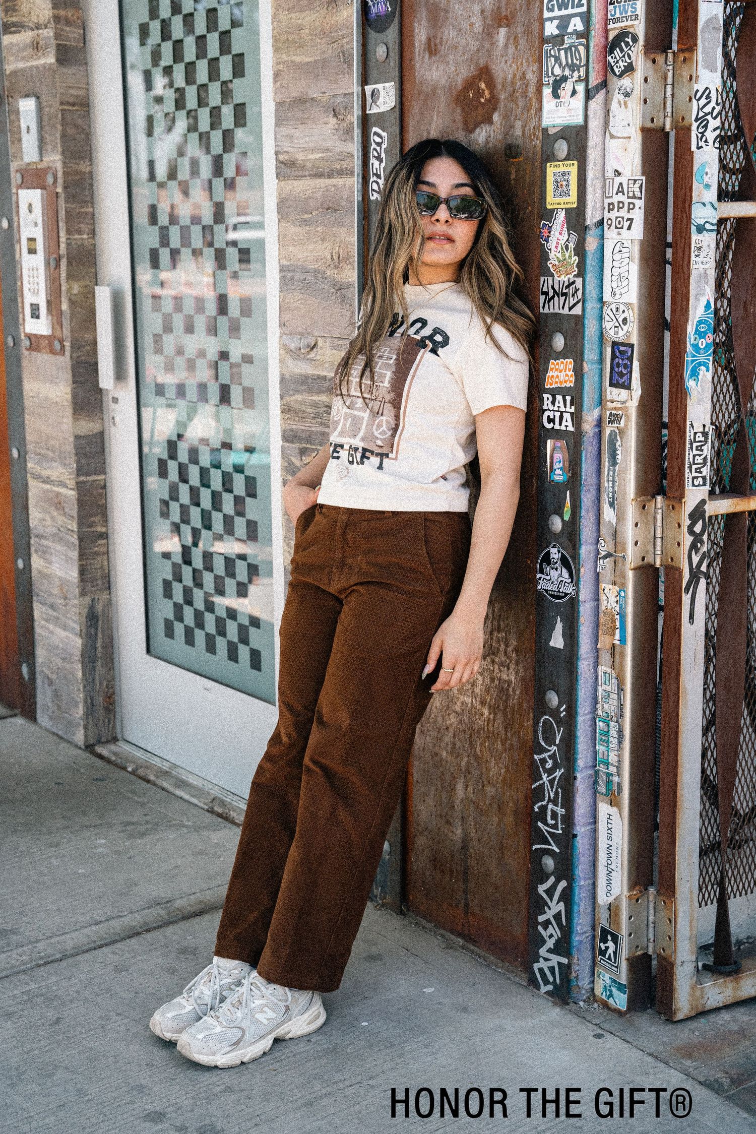 Model wearing brown overalls and white t-shirt poses against urban wall with checkered pattern.