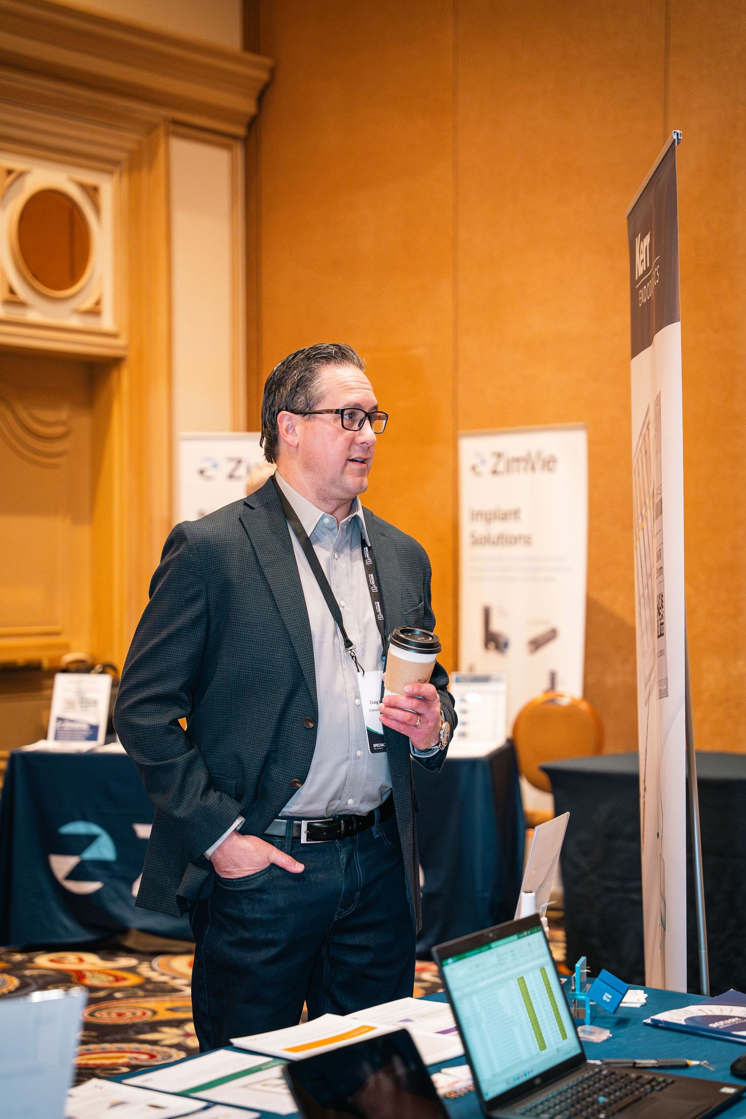 Business professional in suit jacket standing near display booth with laptop at conference.