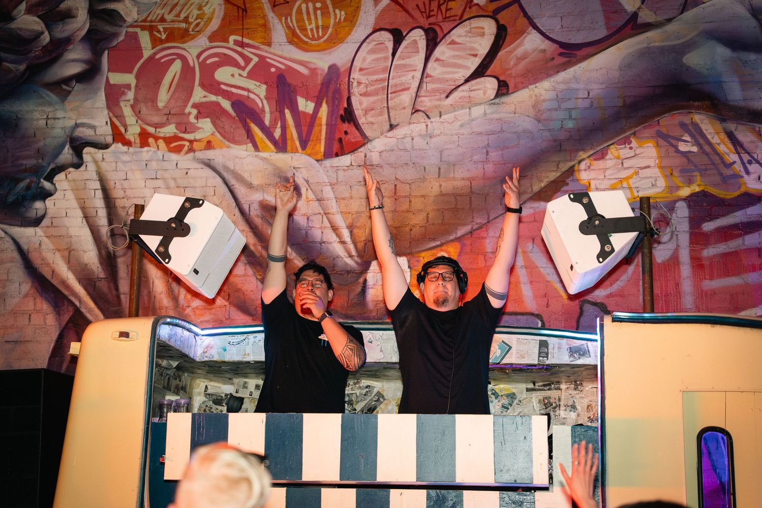 DJ duo performing behind booth with colorful graffiti wall backdrop.
