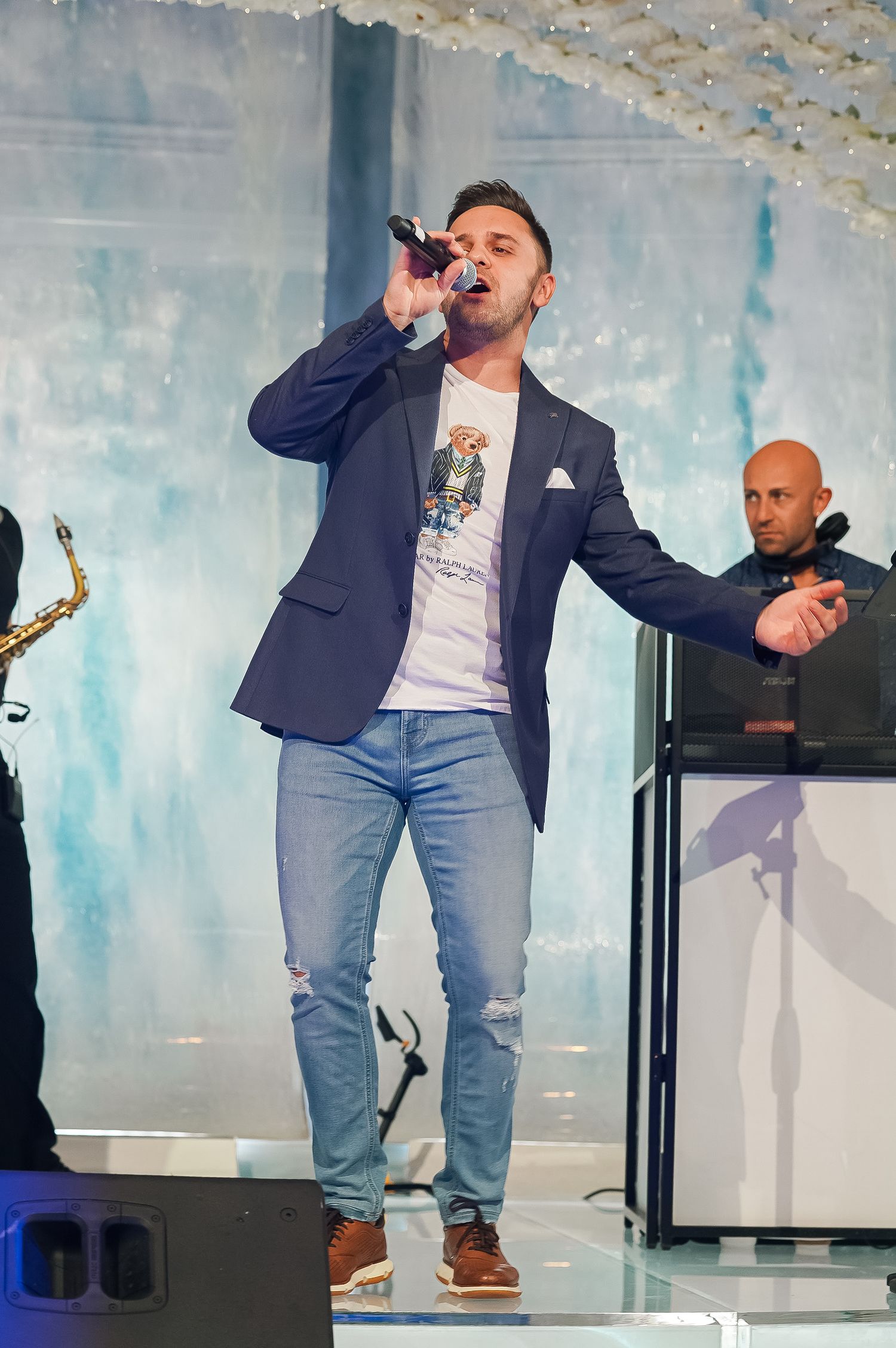 Performer in blue blazer and jeans singing on stage with a microphone.