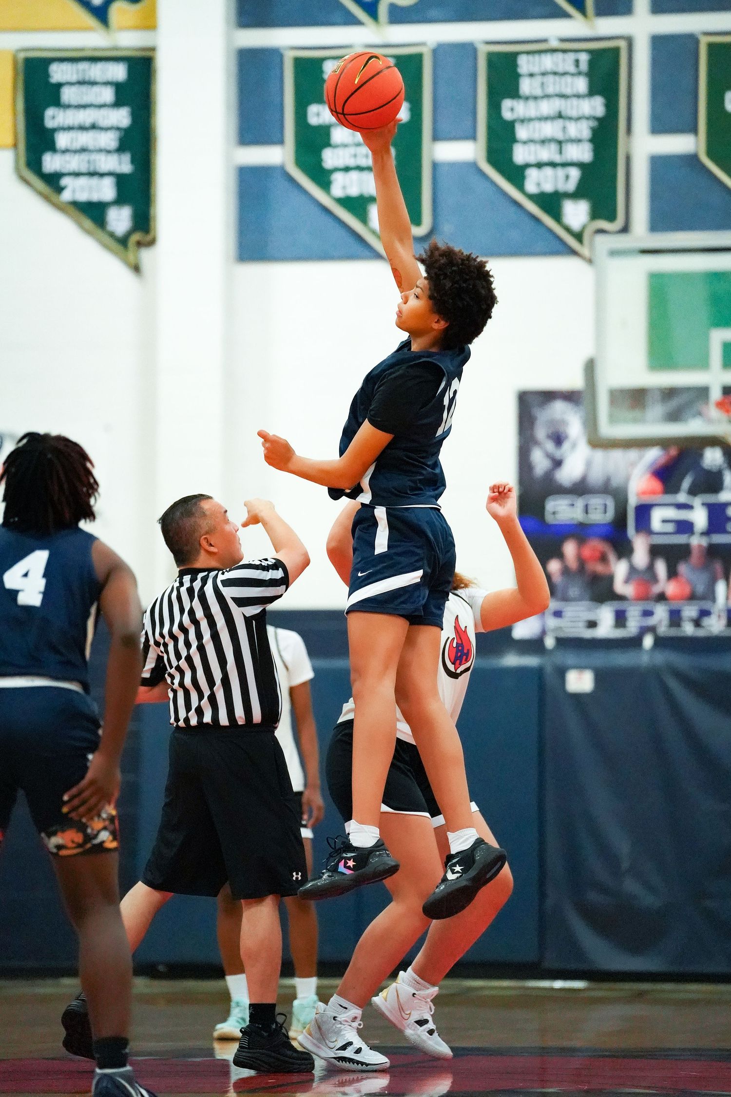 Basketball player leaps high during a jump ball with referee officiating in a high school gymnasium.