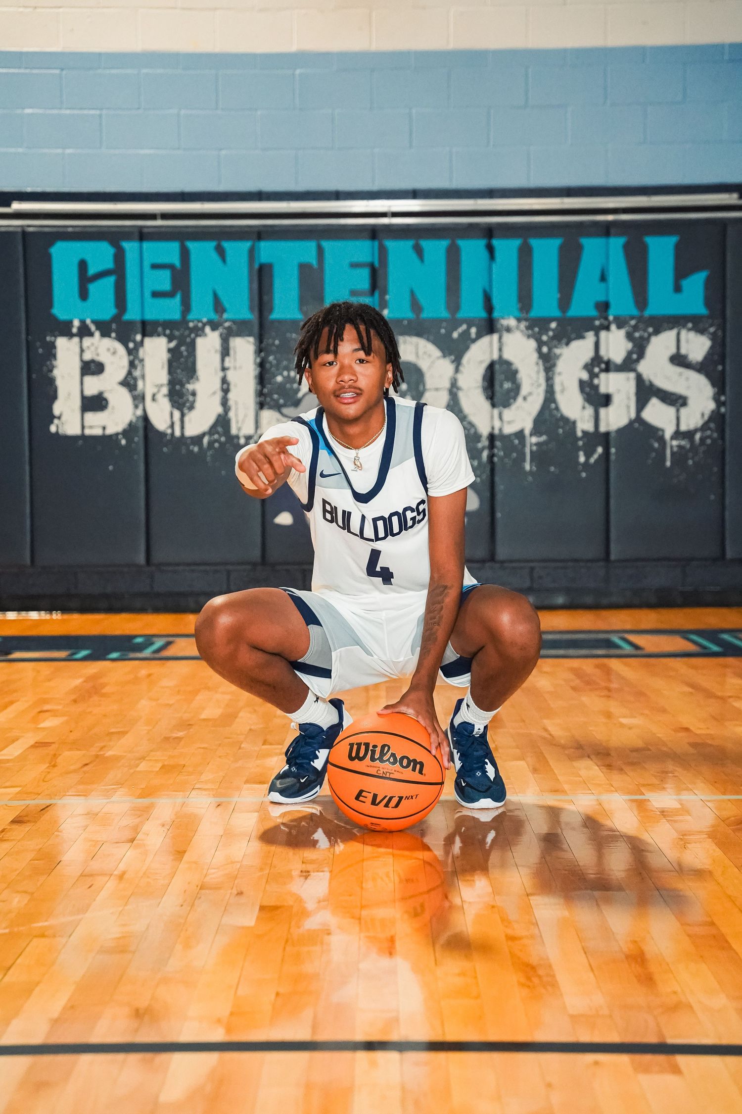 Basketball player in white Bulldogs jersey poses with ball on hardwood court with Centennial wall signage.