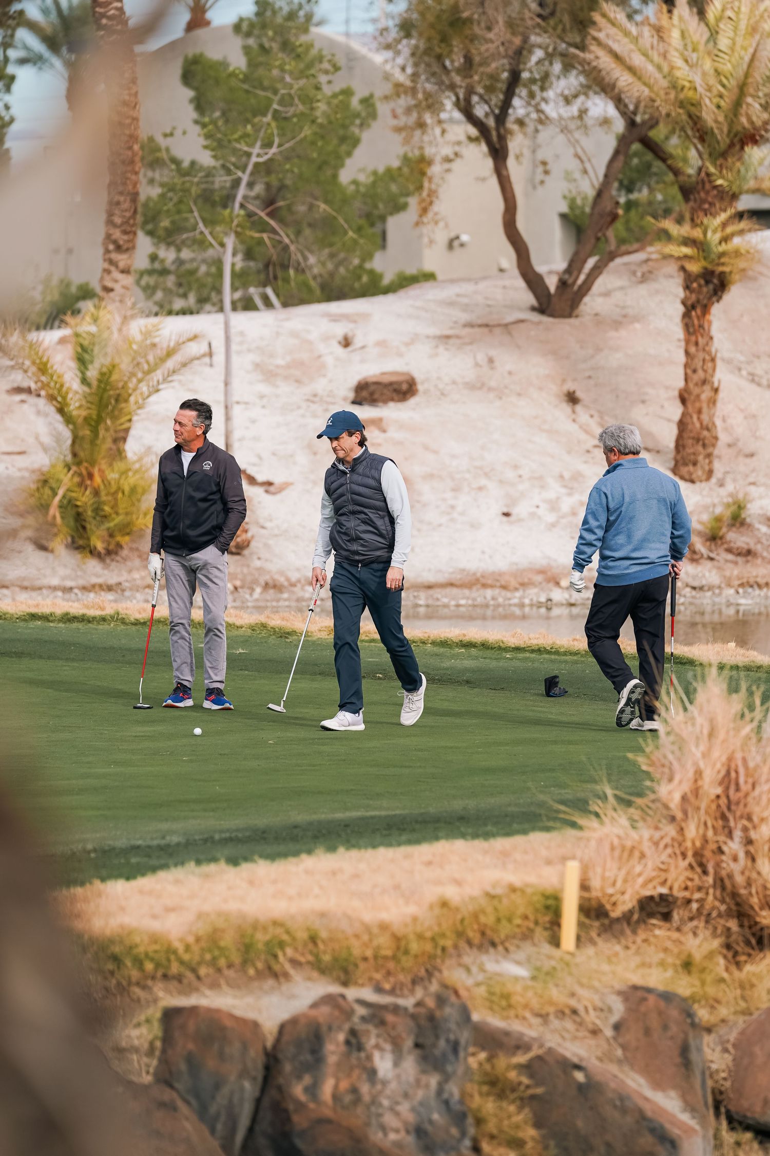 Three golfers stand on a desert course surrounded by palm trees and rocky terrain.