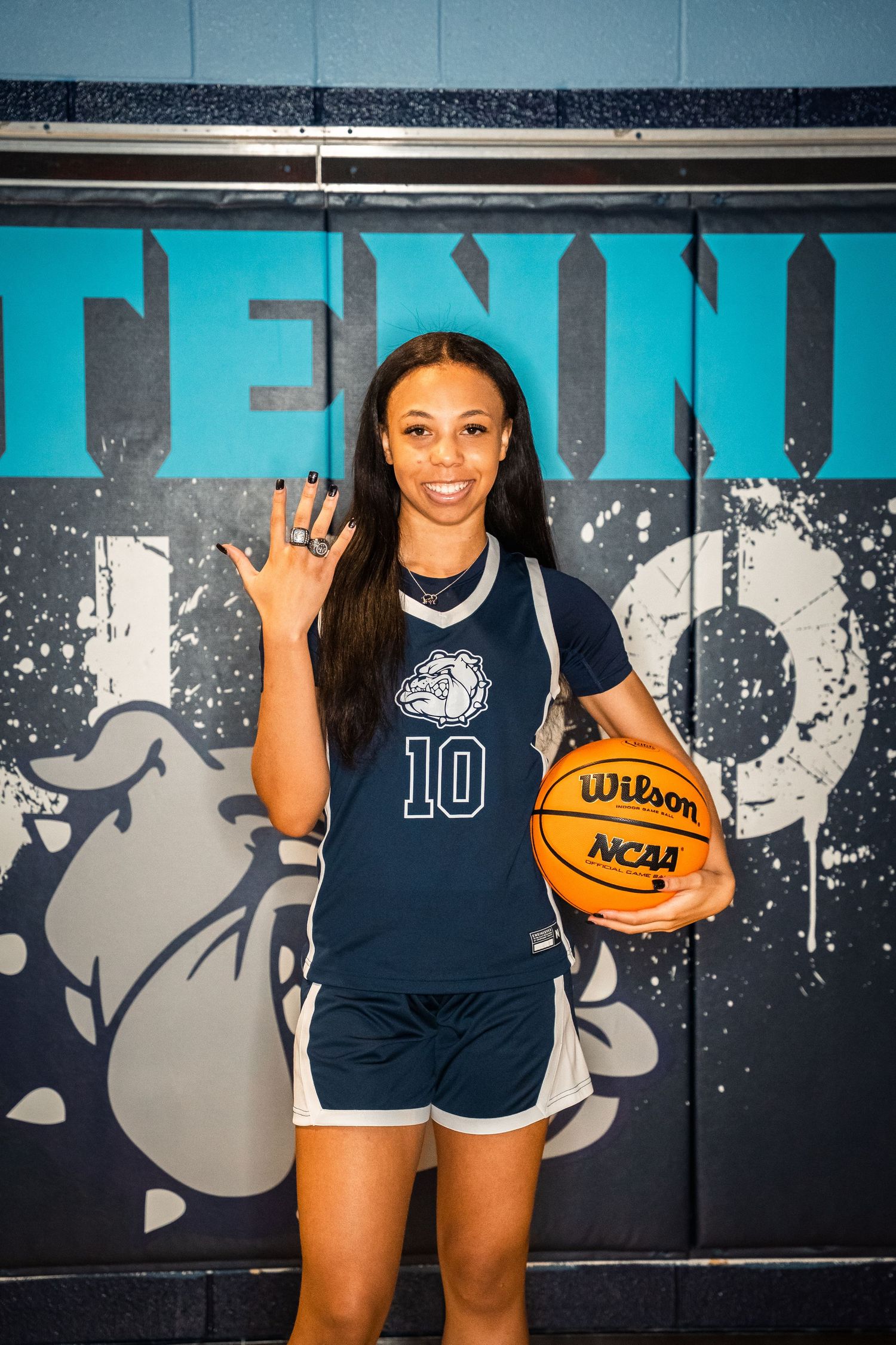 Basketball player posing with Wilson NCAA ball in front of team logo wall.