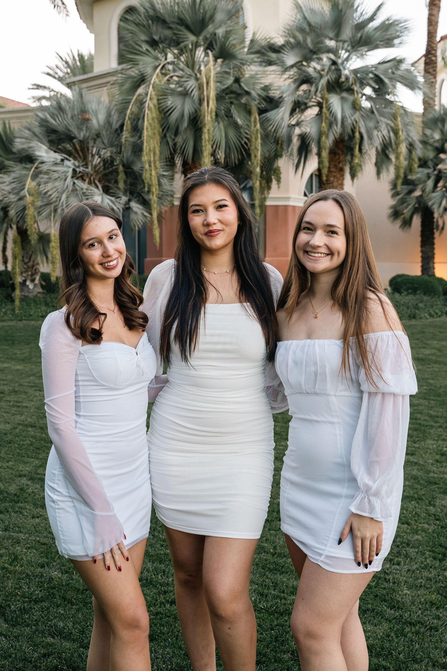 Three friends in white dresses pose together in front of palm trees and Mediterranean architecture.