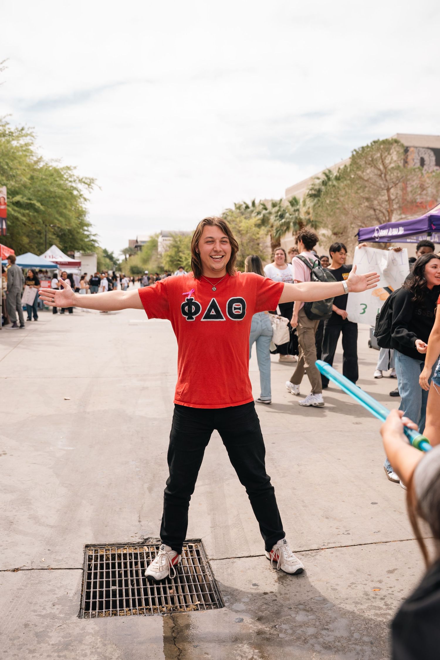 Student in red Greek letter shirt poses with arms spread at campus event.