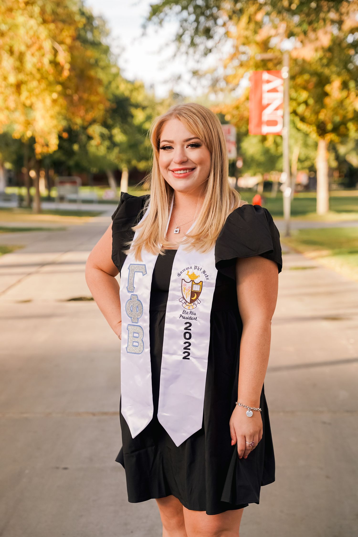 Graduate wearing black dress and white graduation stoles poses on campus sidewalk with autumn trees in background.