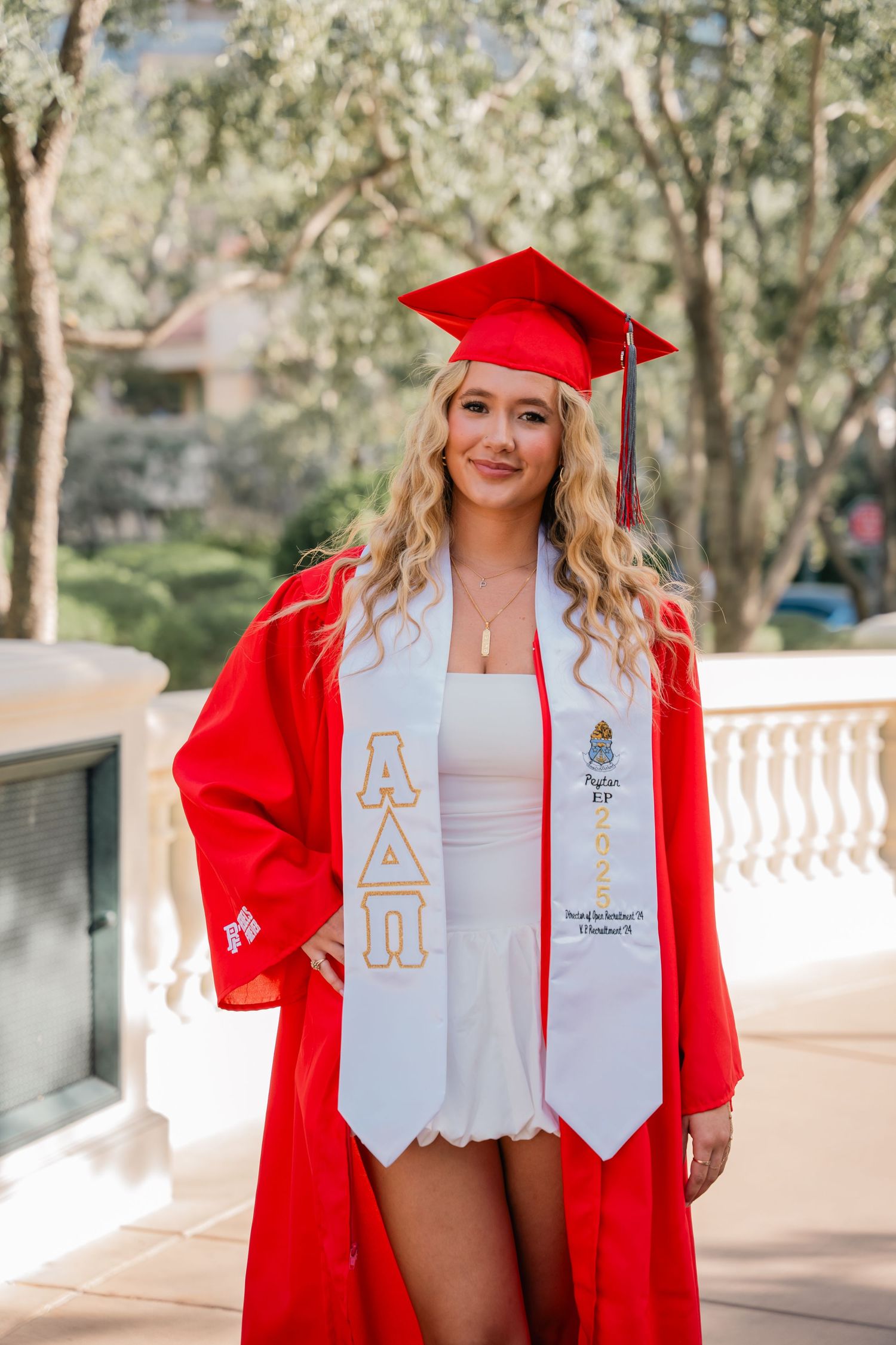 Graduate wearing red cap and gown poses with decorated stoles in outdoor campus setting.