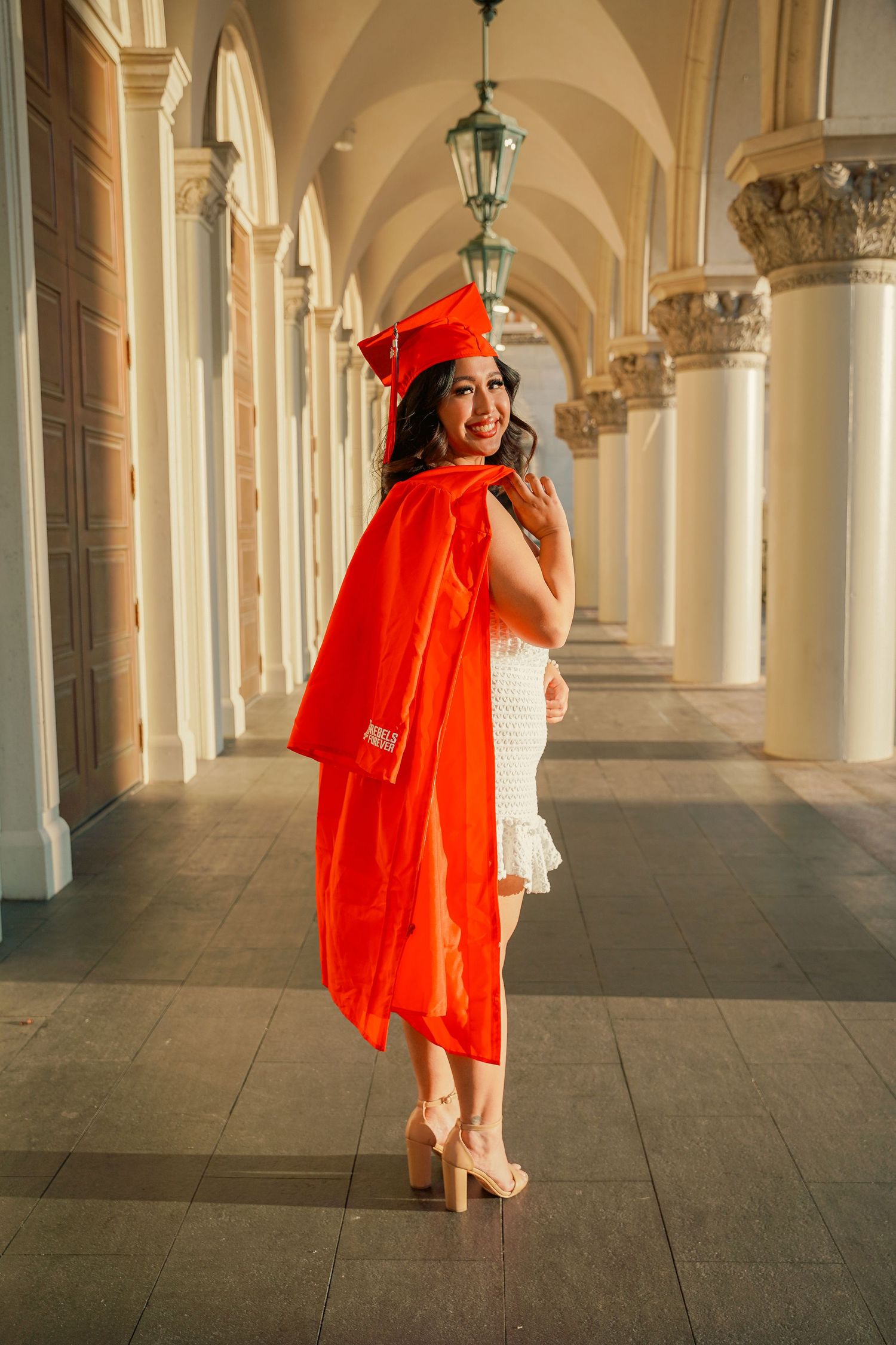 Graduate in orange cap and gown poses elegantly in arched hallway with classical columns.