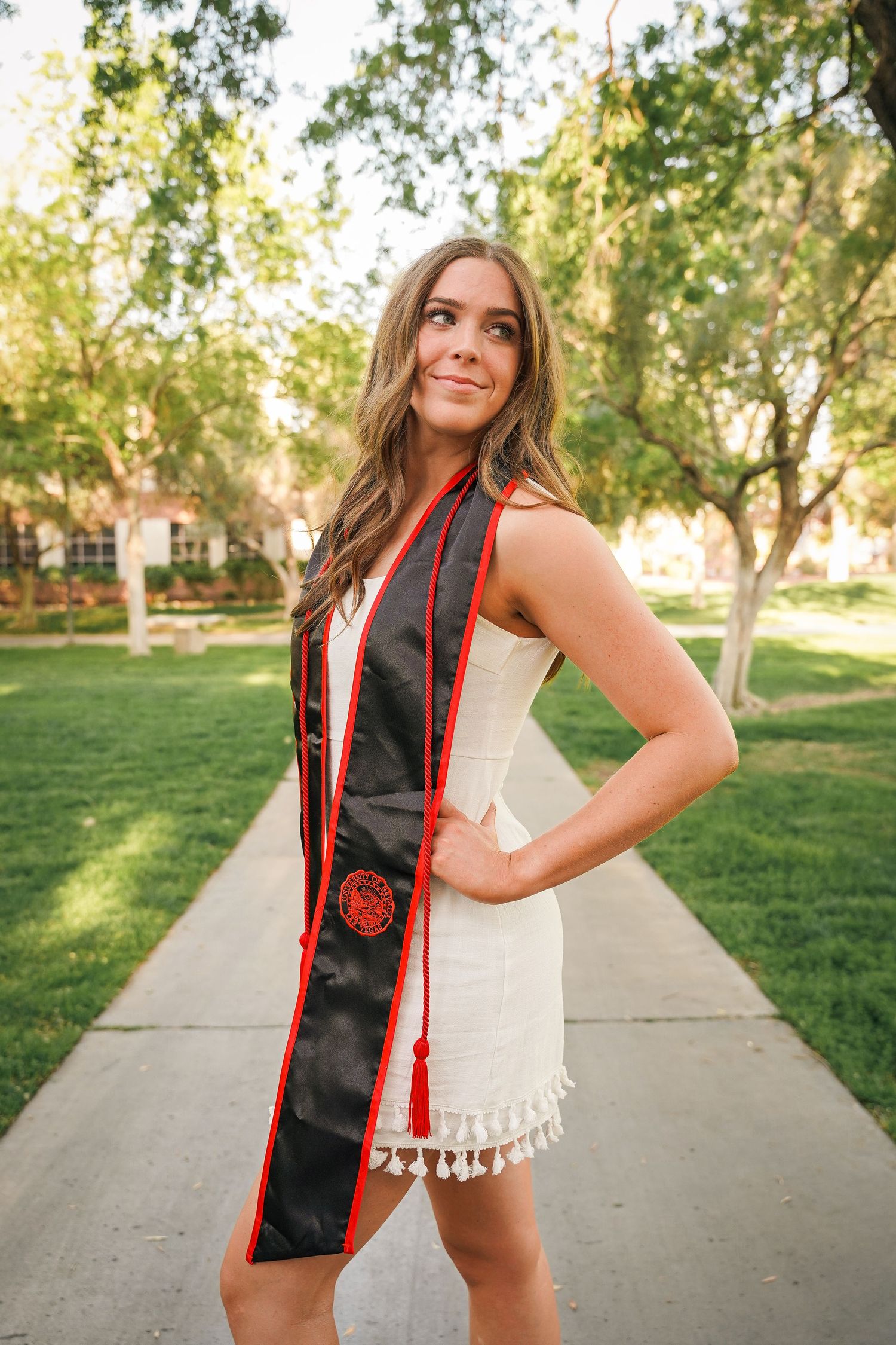 Graduate wearing white dress and graduation sash poses on sunny campus walkway.
