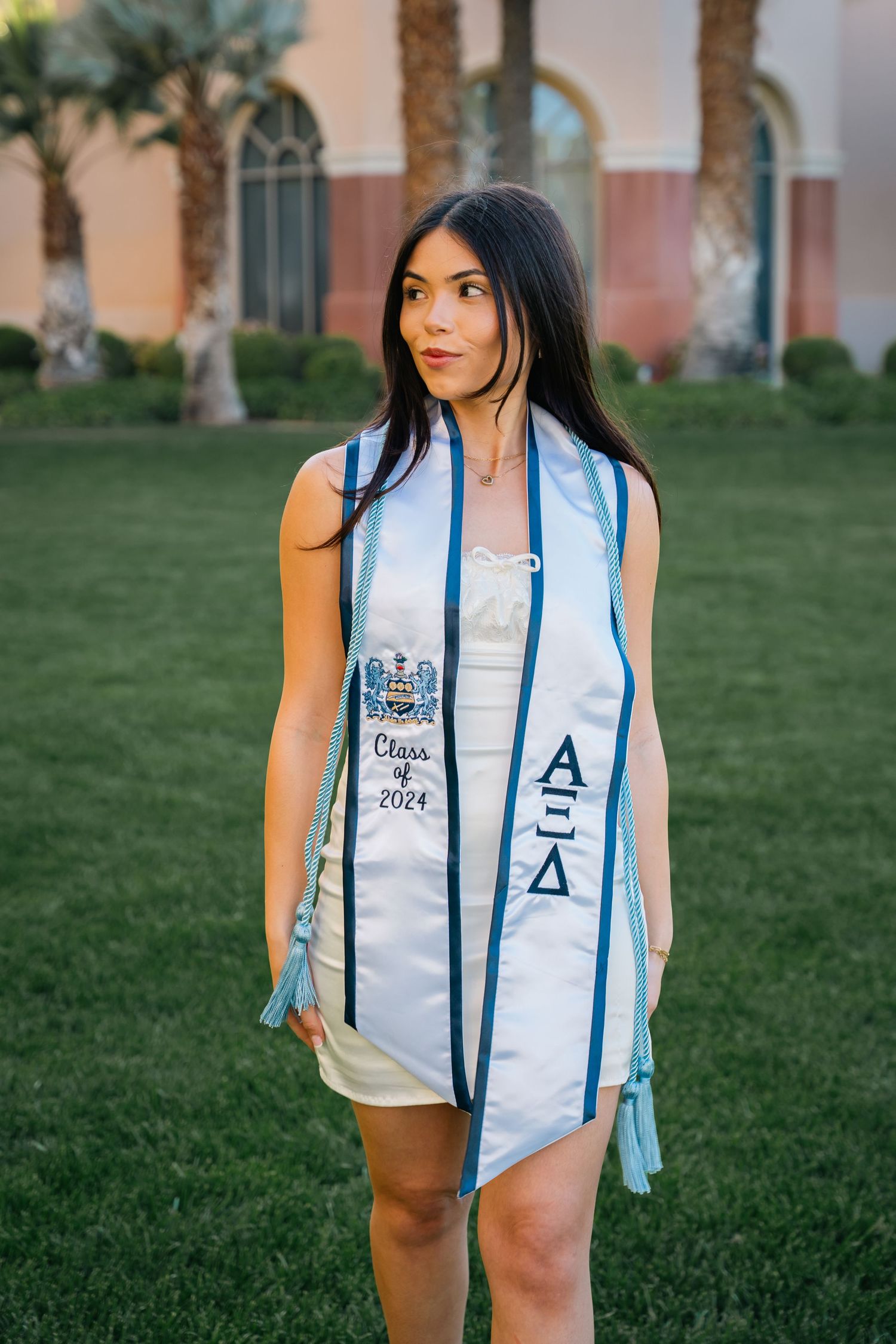 Graduate in white dress with Greek lettered stole poses in front of Mediterranean style building.