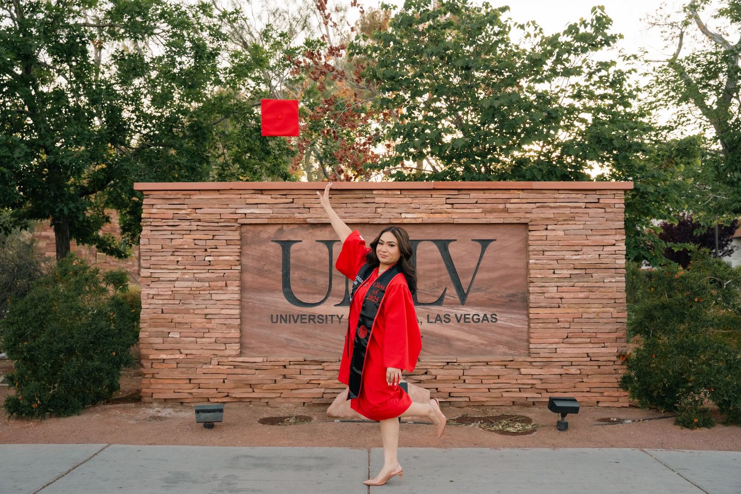 Graduate takes celebratory photos by UNLV stone sign surrounded by desert landscaping.