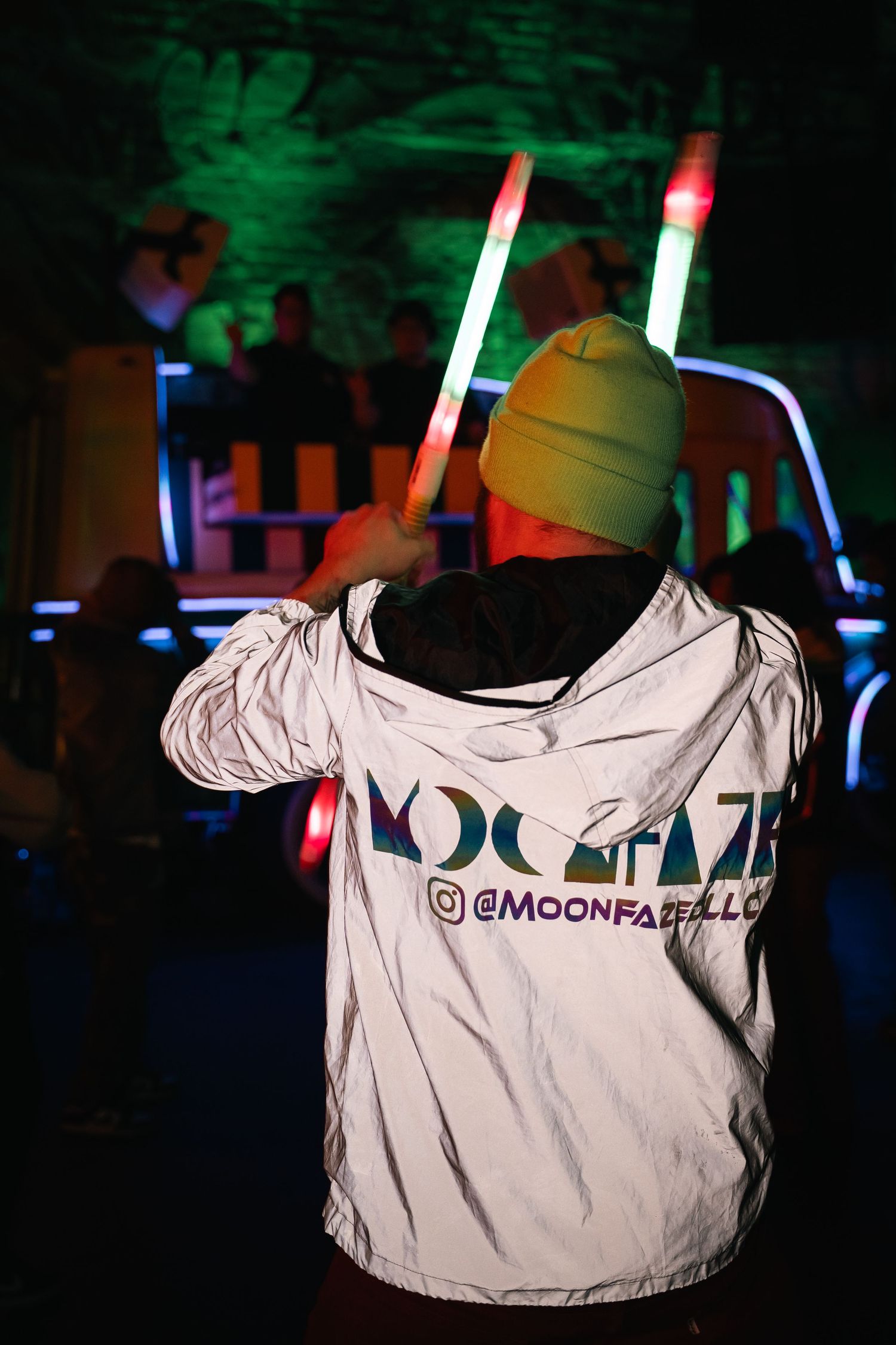 Person wearing yellow beanie and white Moonpath shirt holding light sticks in dark club setting.
