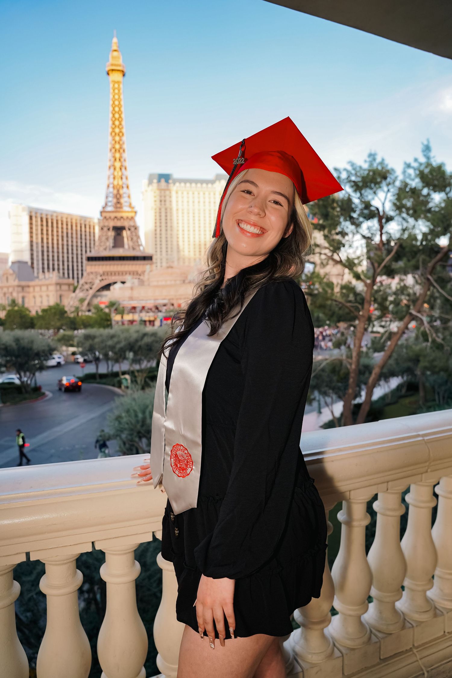 Graduate in cap and gown poses on balcony with Eiffel Tower replica in Las Vegas background.