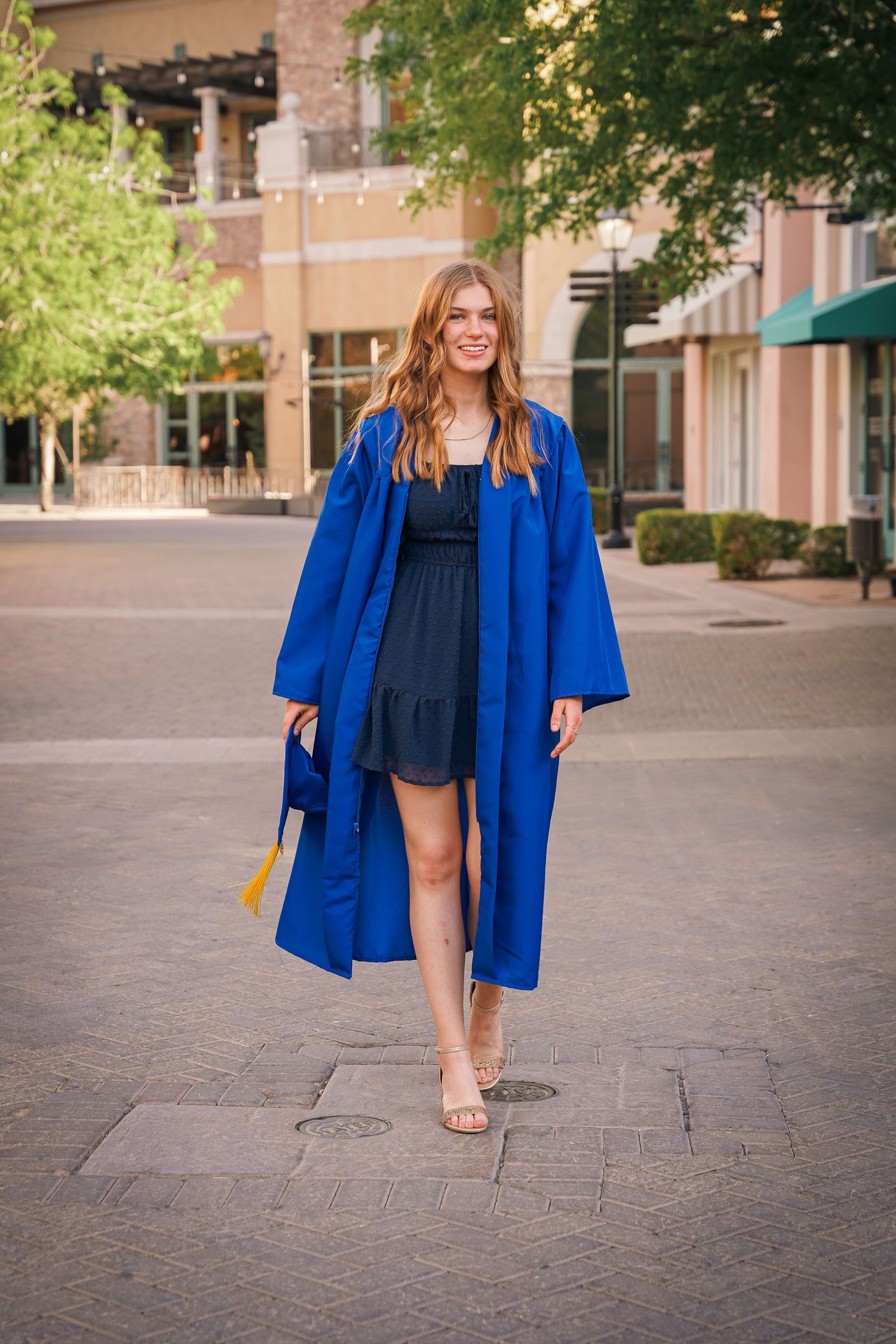 Graduate wearing a royal blue graduation gown over a navy dress walking outdoors on campus.