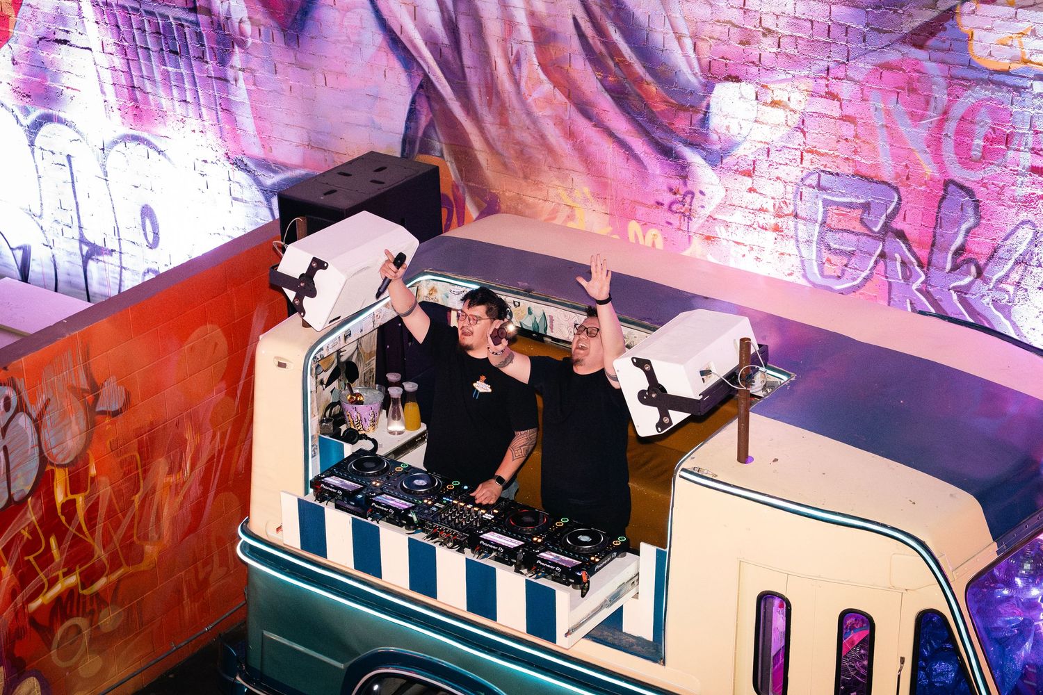 Party crowd enjoying music from custom DJ booth shaped like retro vehicle with pink lighting.