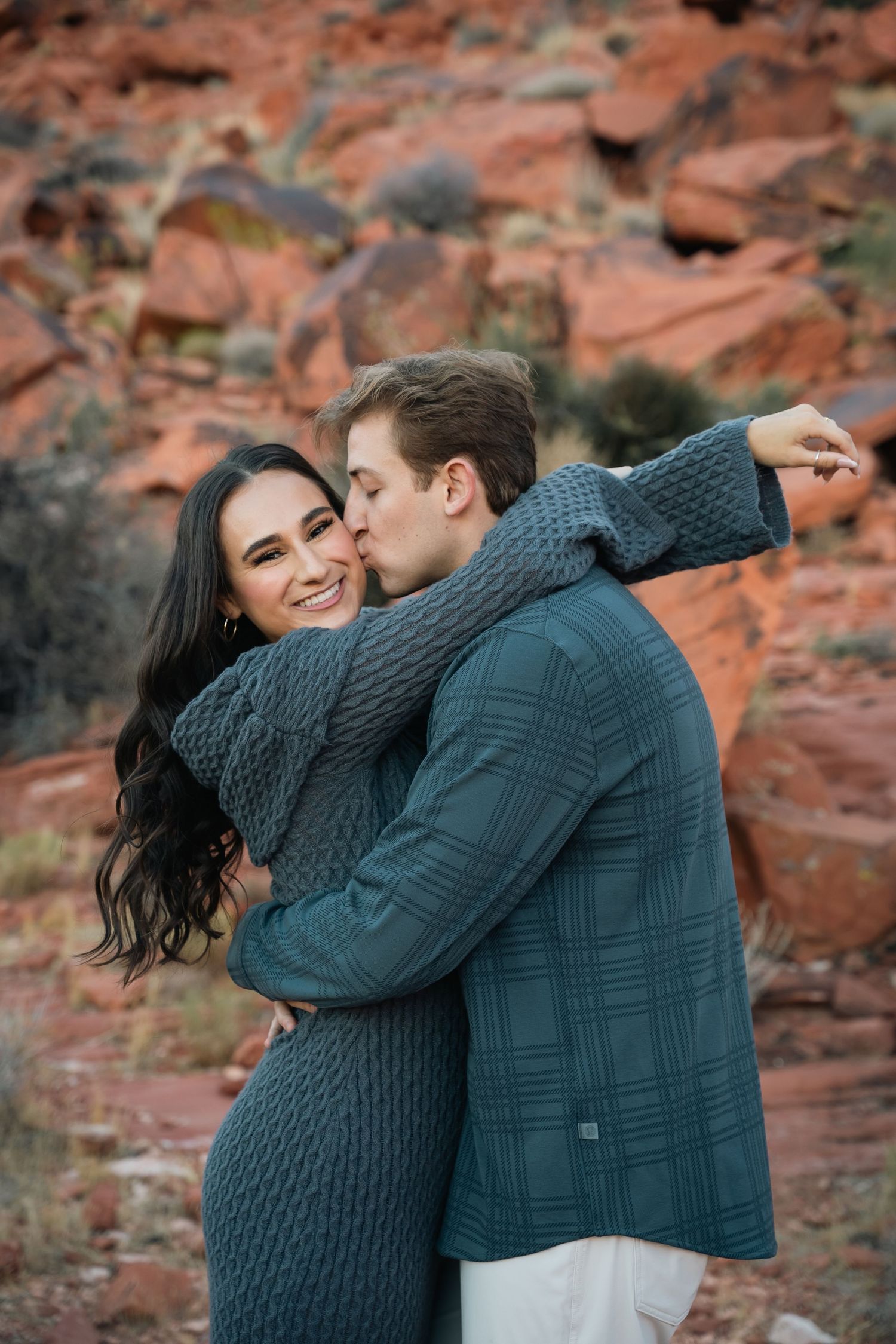 A couple in knit sweaters sharing a playful moment against vibrant red canyon walls.