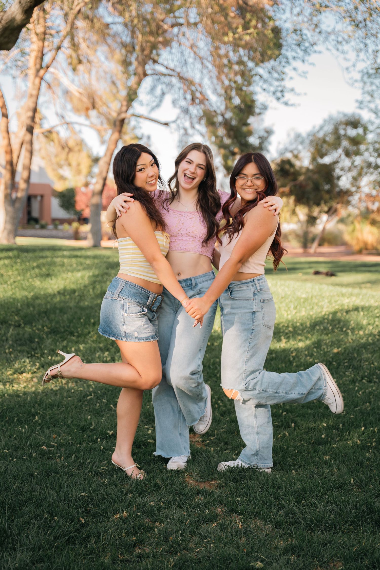 Three friends pose playfully outdoors wearing coordinated outfits of pink tops and denim.