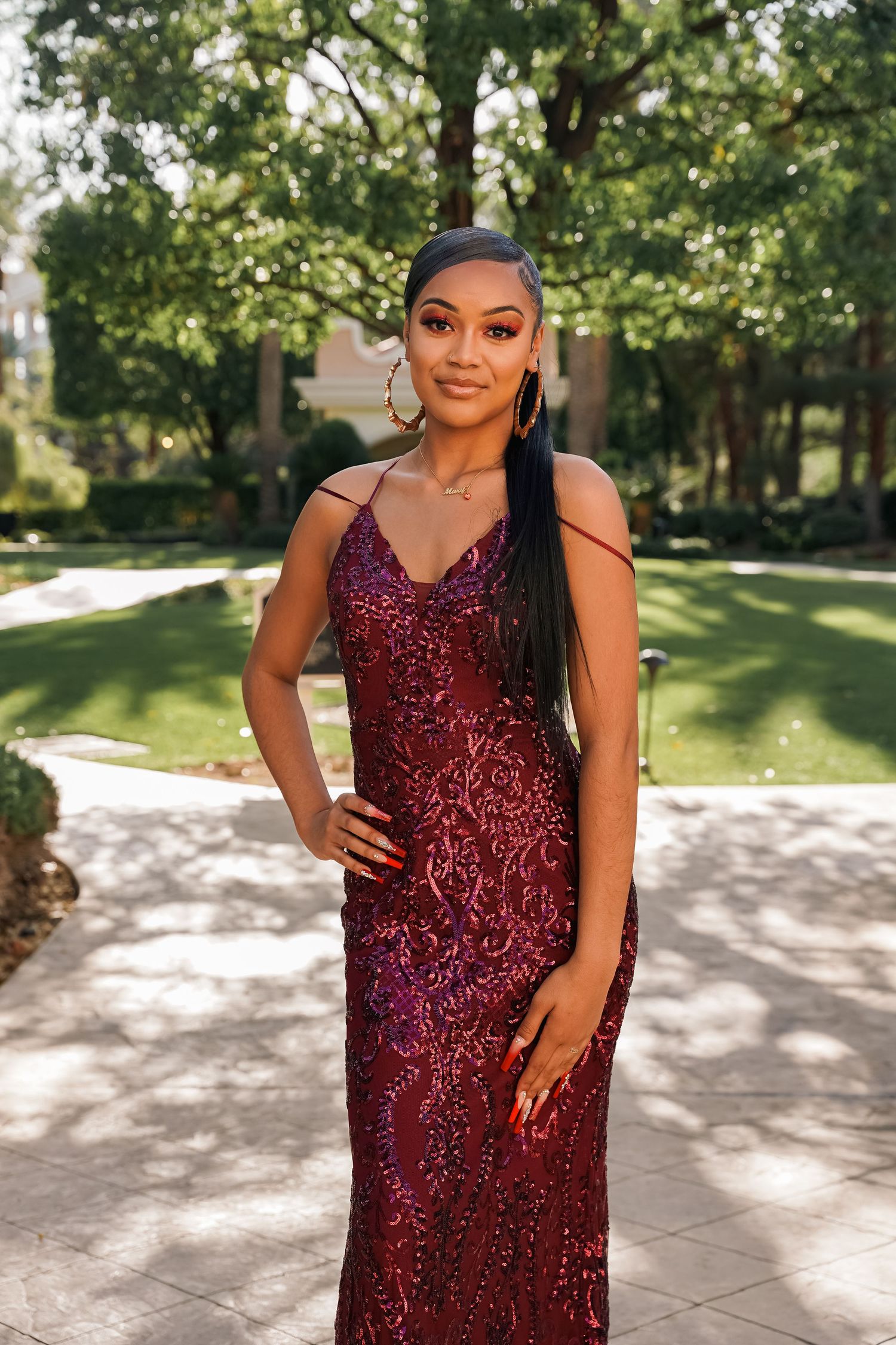 Person in burgundy sequined gown poses on stone pathway surrounded by green trees.