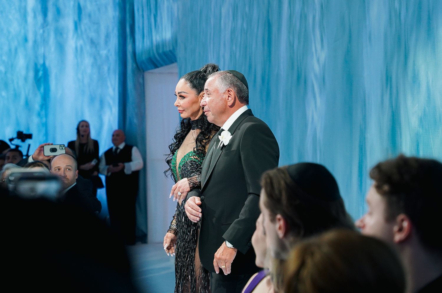 Couple walking down the aisle in elegant attire at a formal event with blue-tinted backdrop.