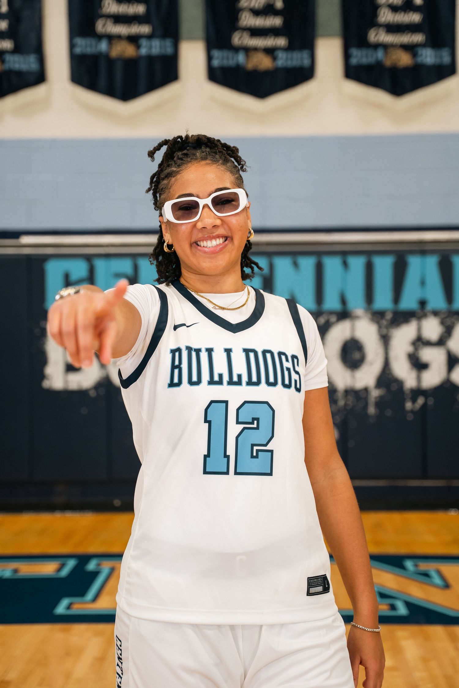 Player in white Bulldogs uniform number 12 with sunglasses points while smiling on basketball court.