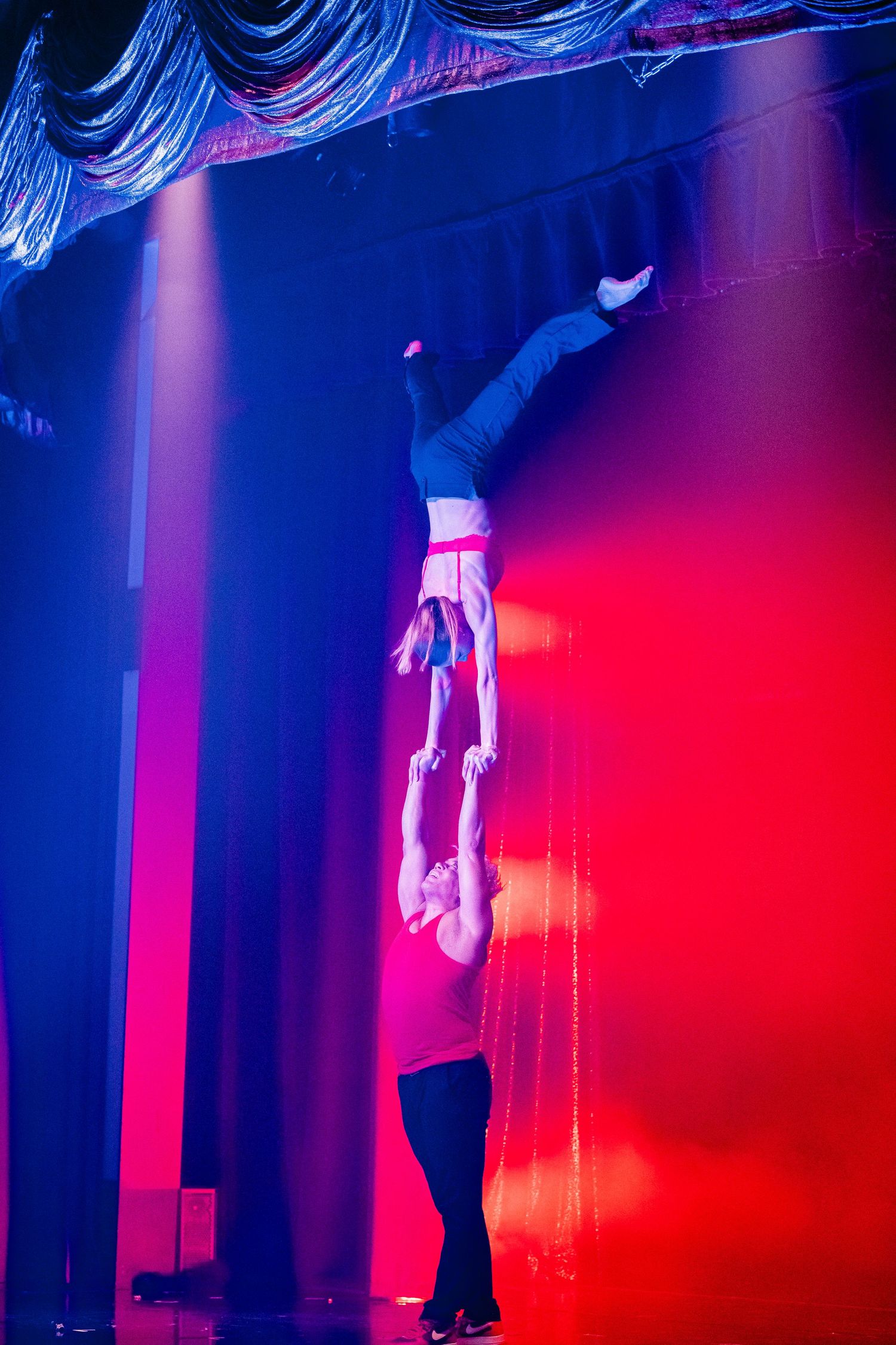 Acrobatic duo performing aerial lift against red and blue stage lighting.
