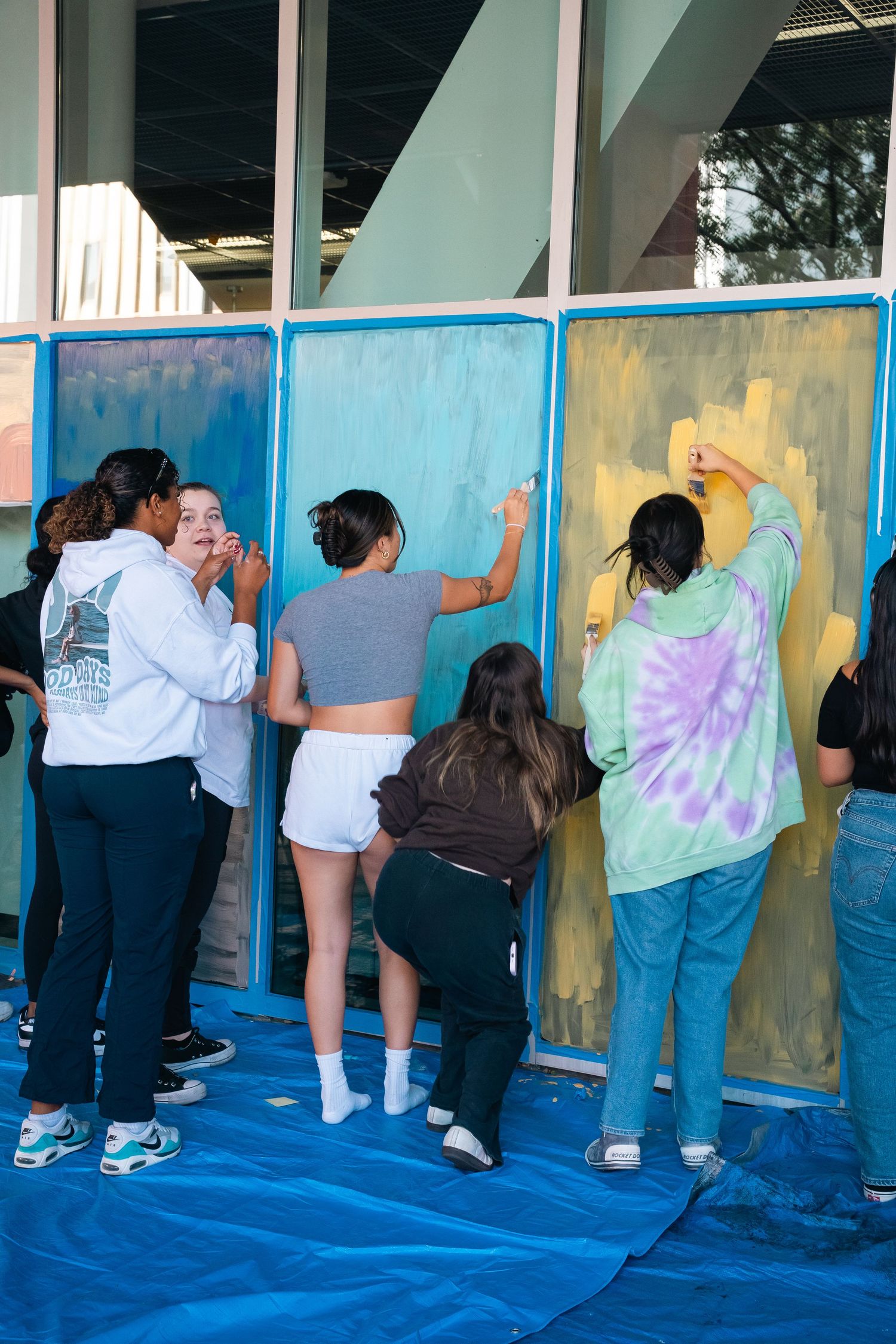 Students painting colorful panels on blue tarp at outdoor event.