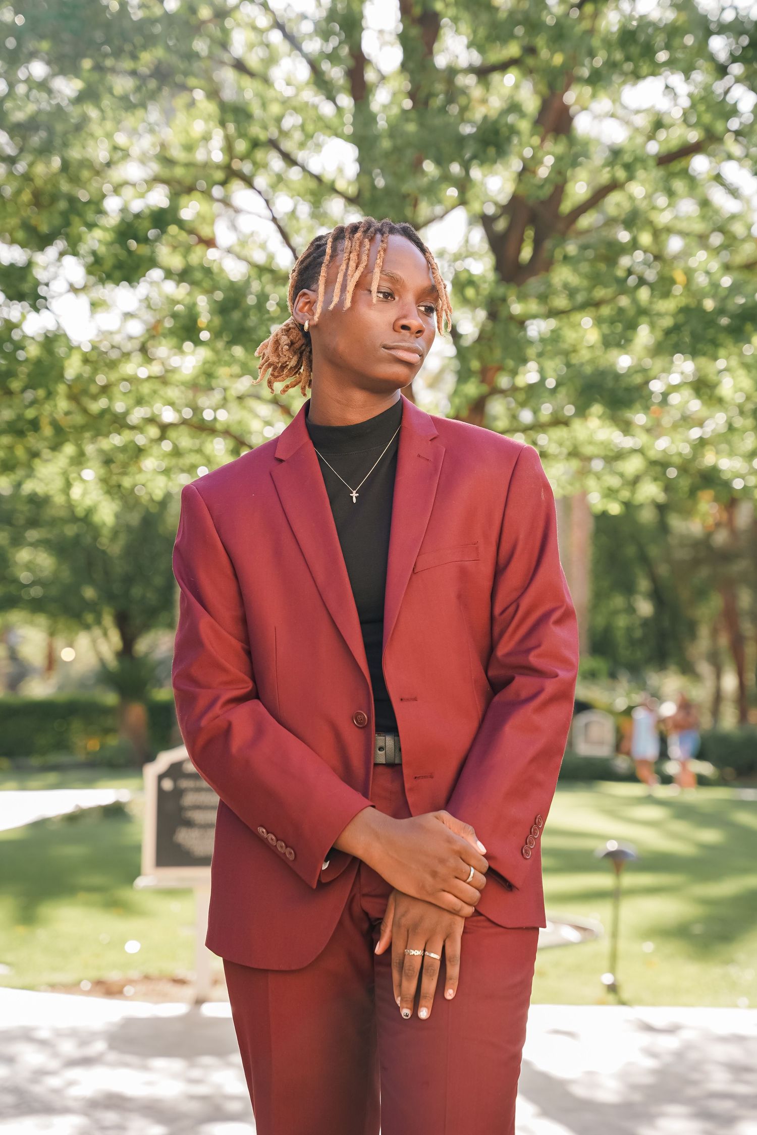 Stylish person poses outdoors in a burgundy suit and black turtleneck against a sunlit garden background.