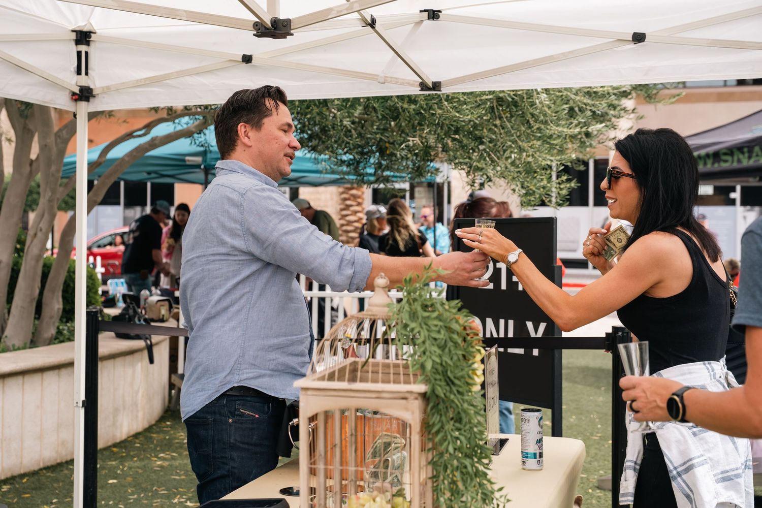 Vendor serving drinks to customers under white tent at outdoor festival.