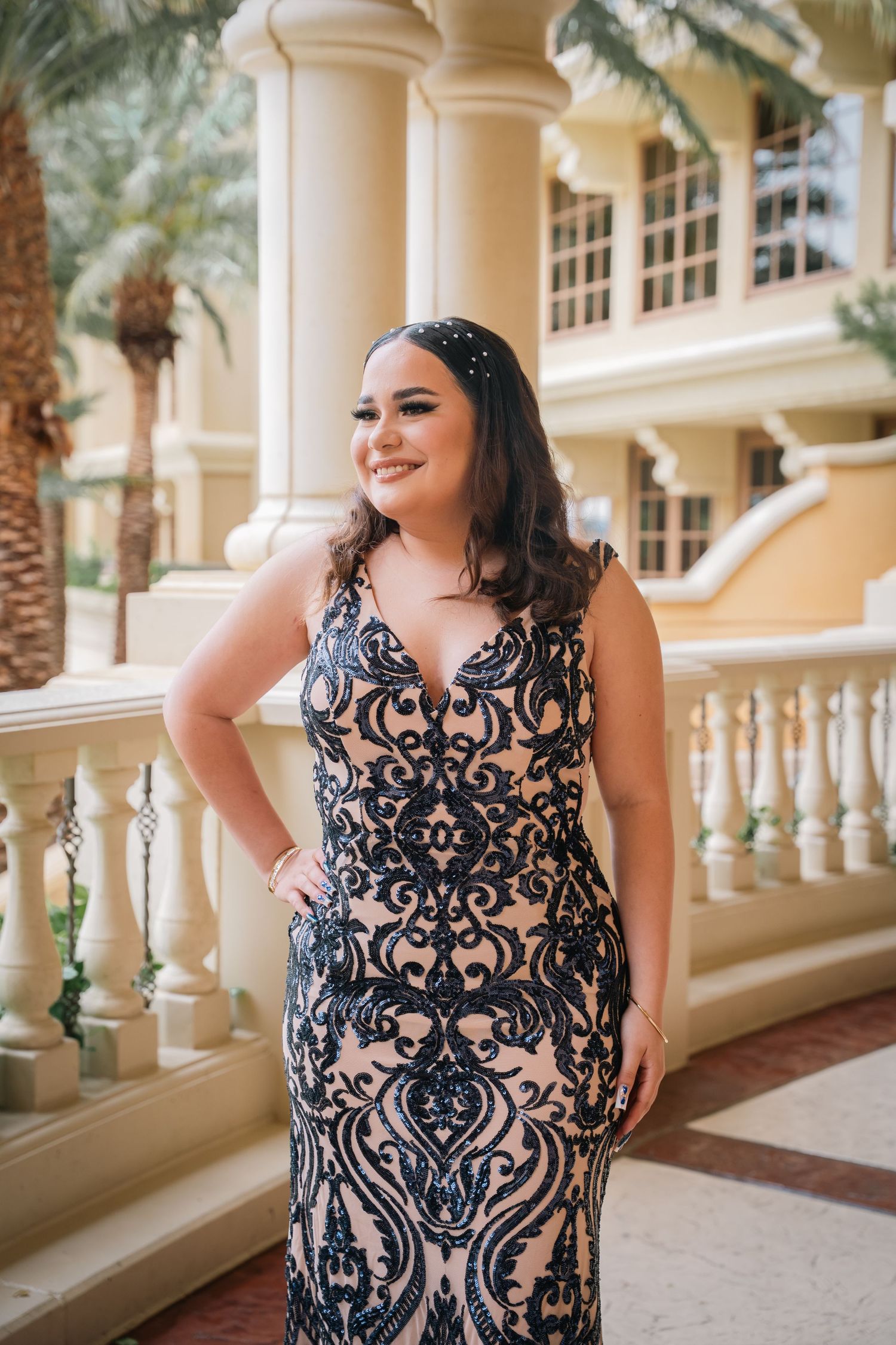 A person in an ornate black and nude lace gown poses on a balcony with Mediterranean-style architecture.