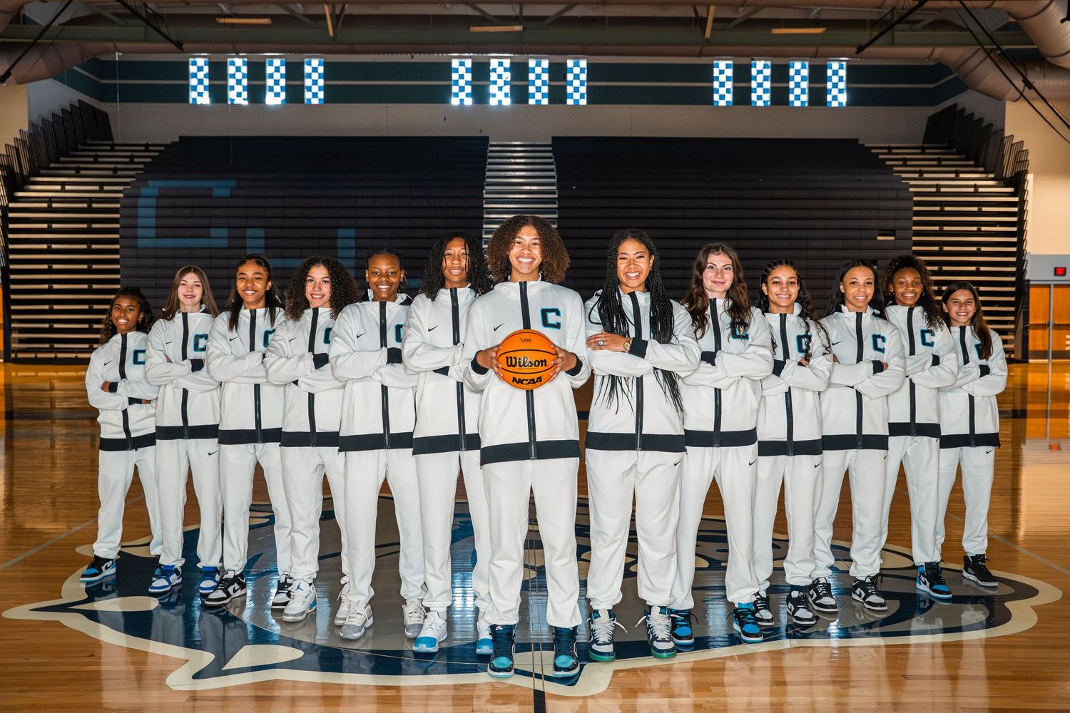 High school basketball team poses in white uniforms on court with mascot logo for team photo.