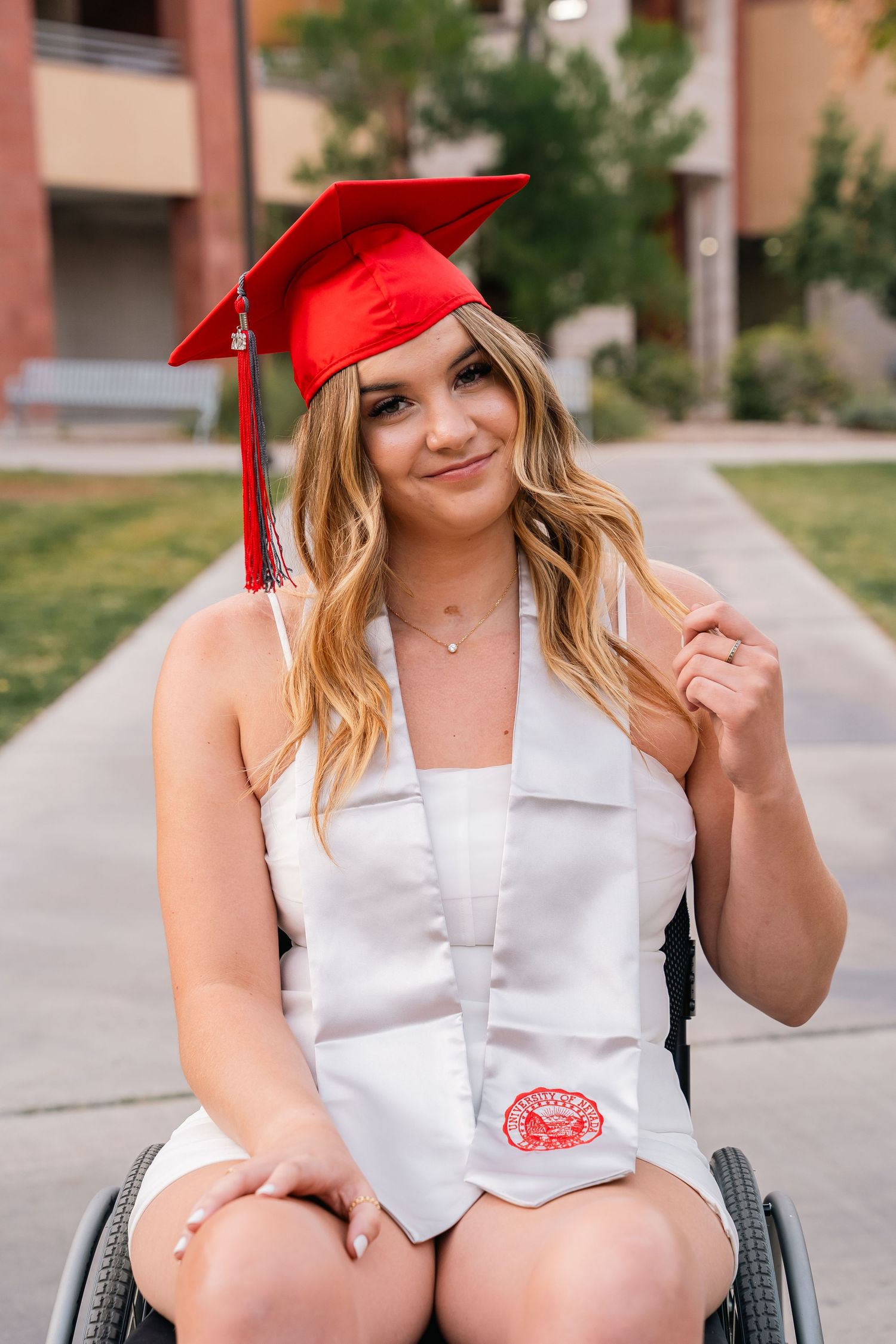 Graduate in wheelchair wearing white dress and red graduation cap smiling on campus pathway.