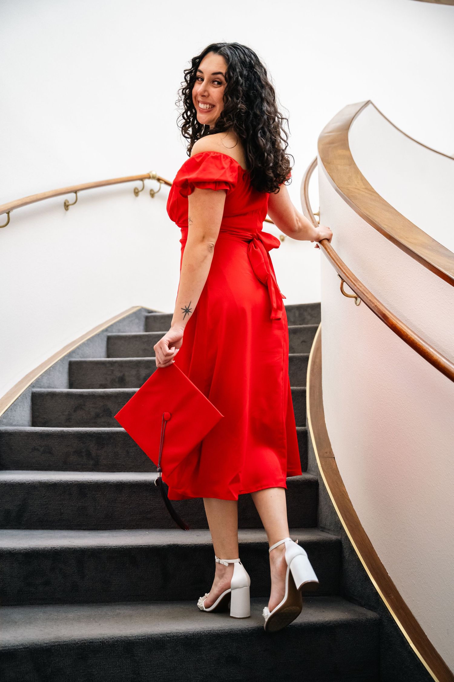 Graduate in red dress poses elegantly on white staircase during indoor photoshoot.